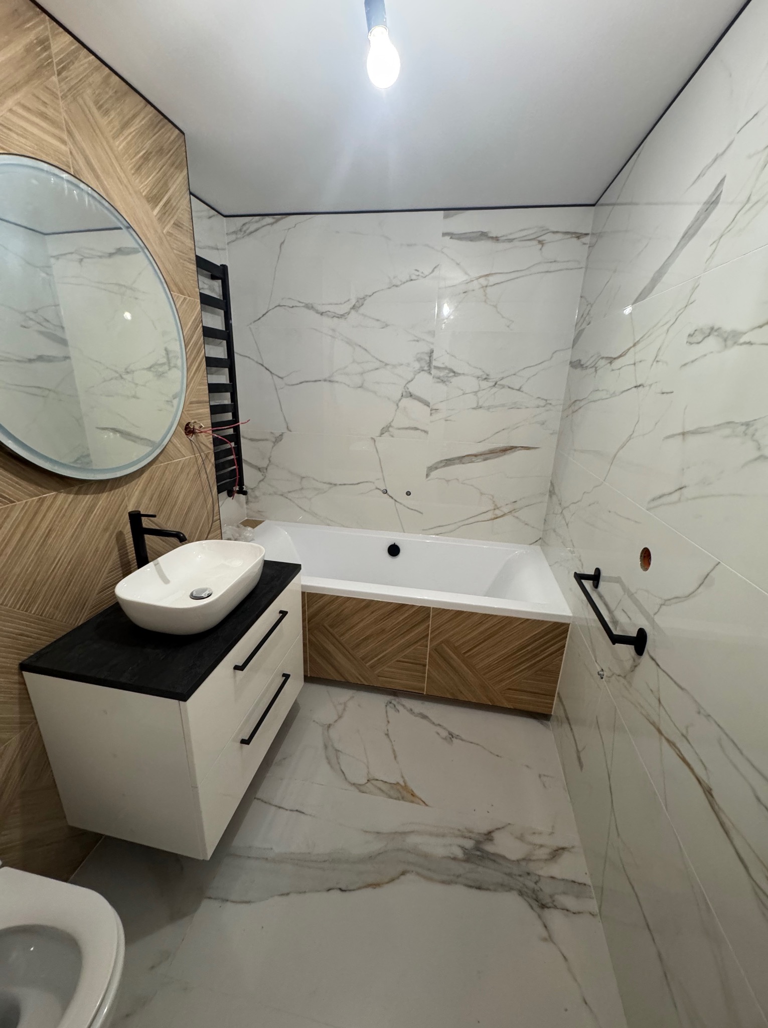 Nowoczesna łazienka z płytkami imitującymi marmur i drewno, owalnym lustrem, białą umywalką nablatową i grzejnikiem drabinkowym.