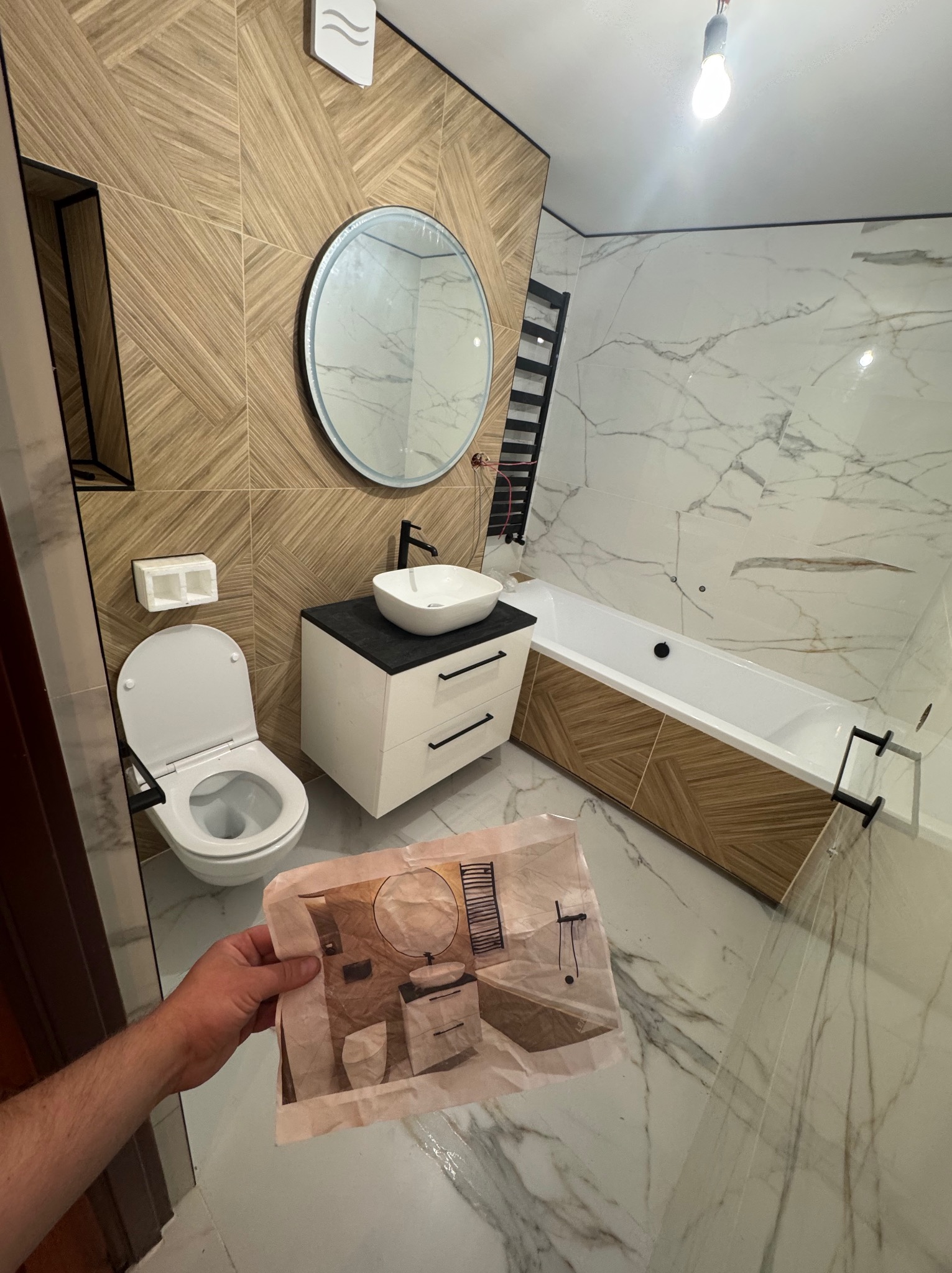 Nowoczesna łazienka z drewnopodobnymi i marmurowymi płytkami, owalnym lustrem i białą armaturą. W dłoni trzymane zdjęcie tej samej łazienki.