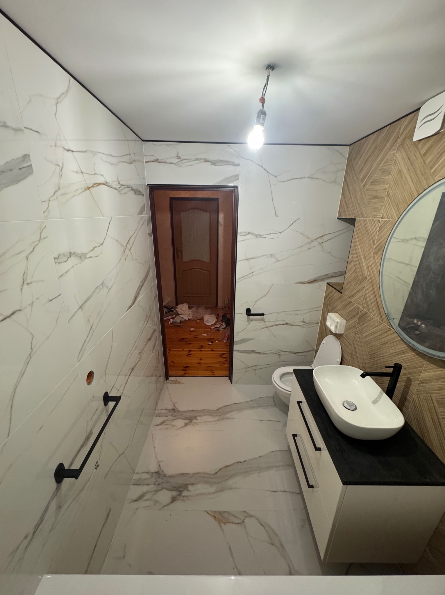 Nowoczesna łazienka z marmurowymi płytkami, drewnianymi akcentami i owalną umywalką na szafce. Widoczna niedokończona podłoga i wisząca żarówka.