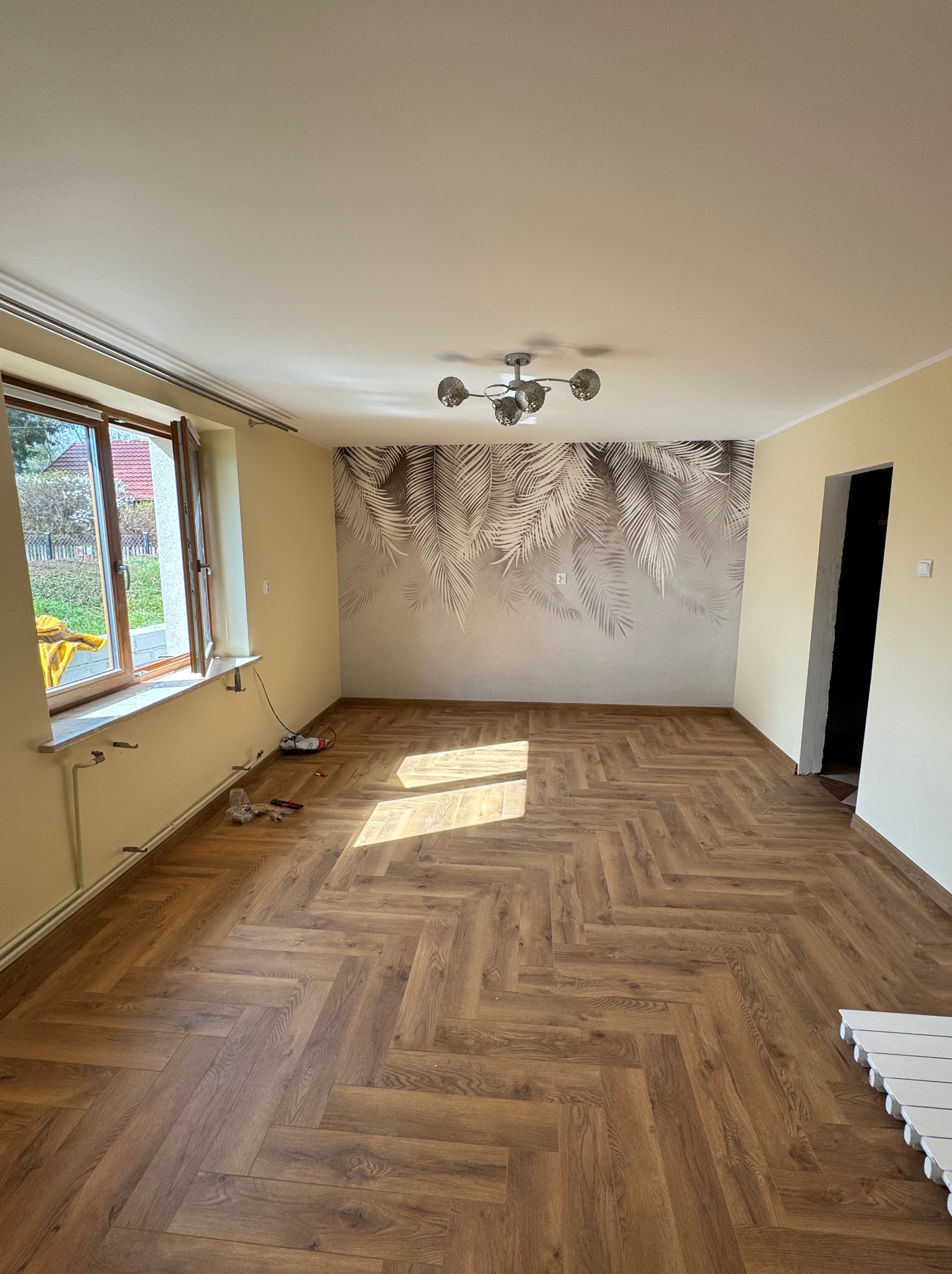 Jasne wnętrze po remoncie: parkiet ułożony w jodełkę, ściany w odcieniach beżu, tapeta z motywem liści, nowoczesna lampa. Widoczne narzędzia przy oknie.