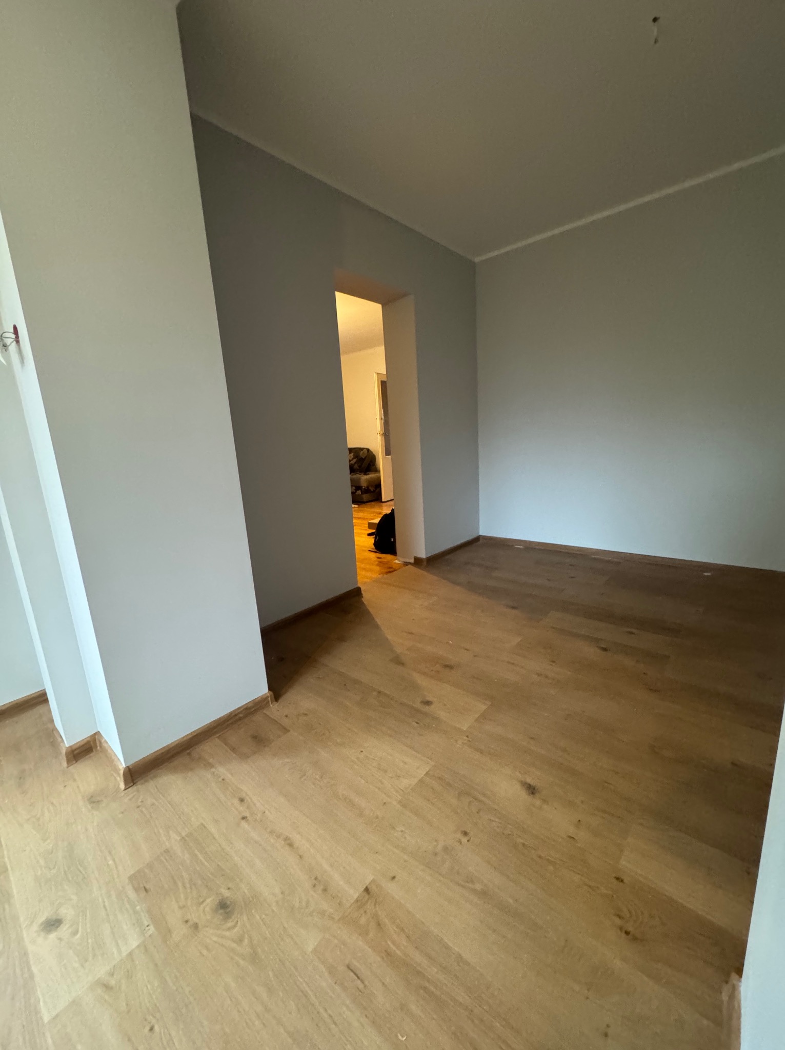 Jasne, drewniane panele podłogowe w pustym pokoju z szarymi ścianami. Widoczne listwy przypodłogowe i otwarte drzwi do kolejnego pomieszczenia.