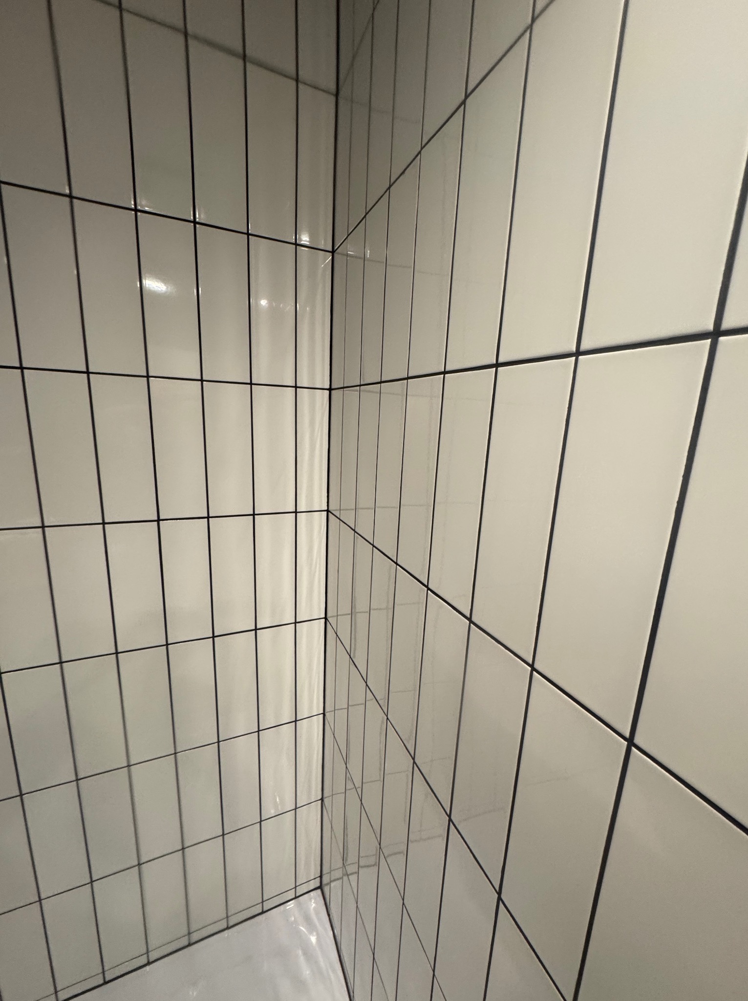 Białe, błyszczące, prostokątne płytki ułożone pionowo z ciemną fugą na ścianach w rogu łazienki. Widoczny fragment brodzika.