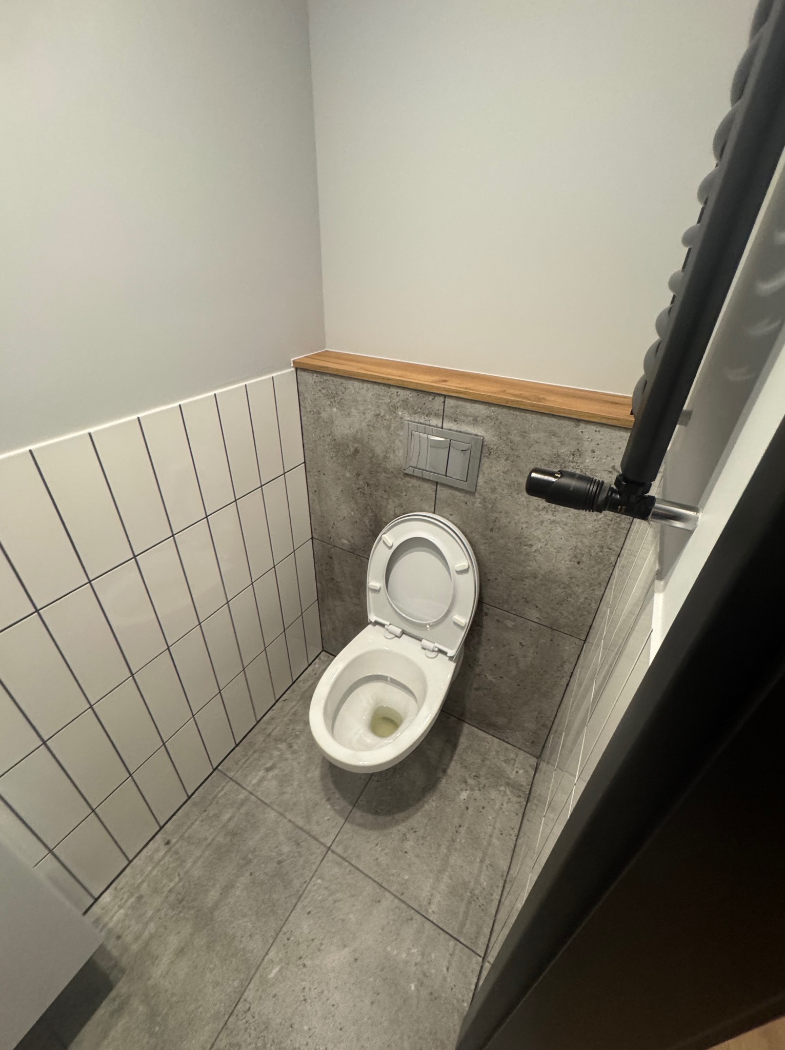 Minimalistyczna toaleta z wiszącą muszlą, szarymi płytkami i białymi, pionowo ułożonymi kafelkami. Drewniany akcent nad spłuczką. Widok z góry.