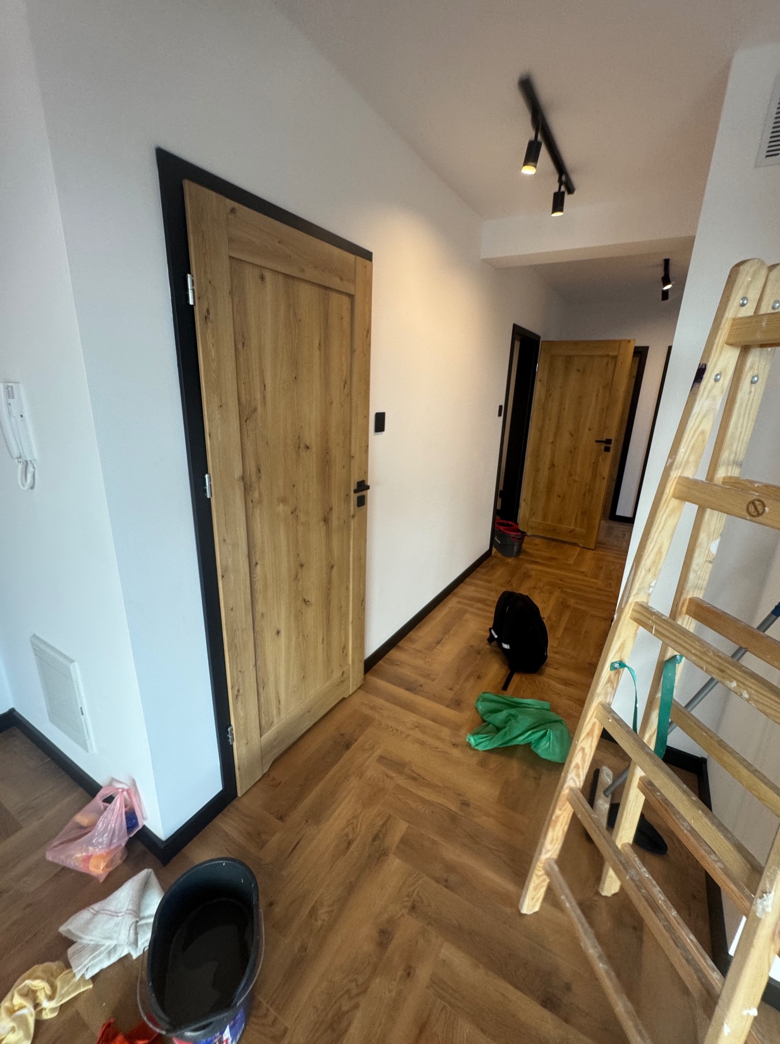 Korytarz po remoncie z drewnianą podłogą w jodełkę, drewnianymi drzwiami z czarnymi ościeżnicami i rozstawioną drabiną malarską.