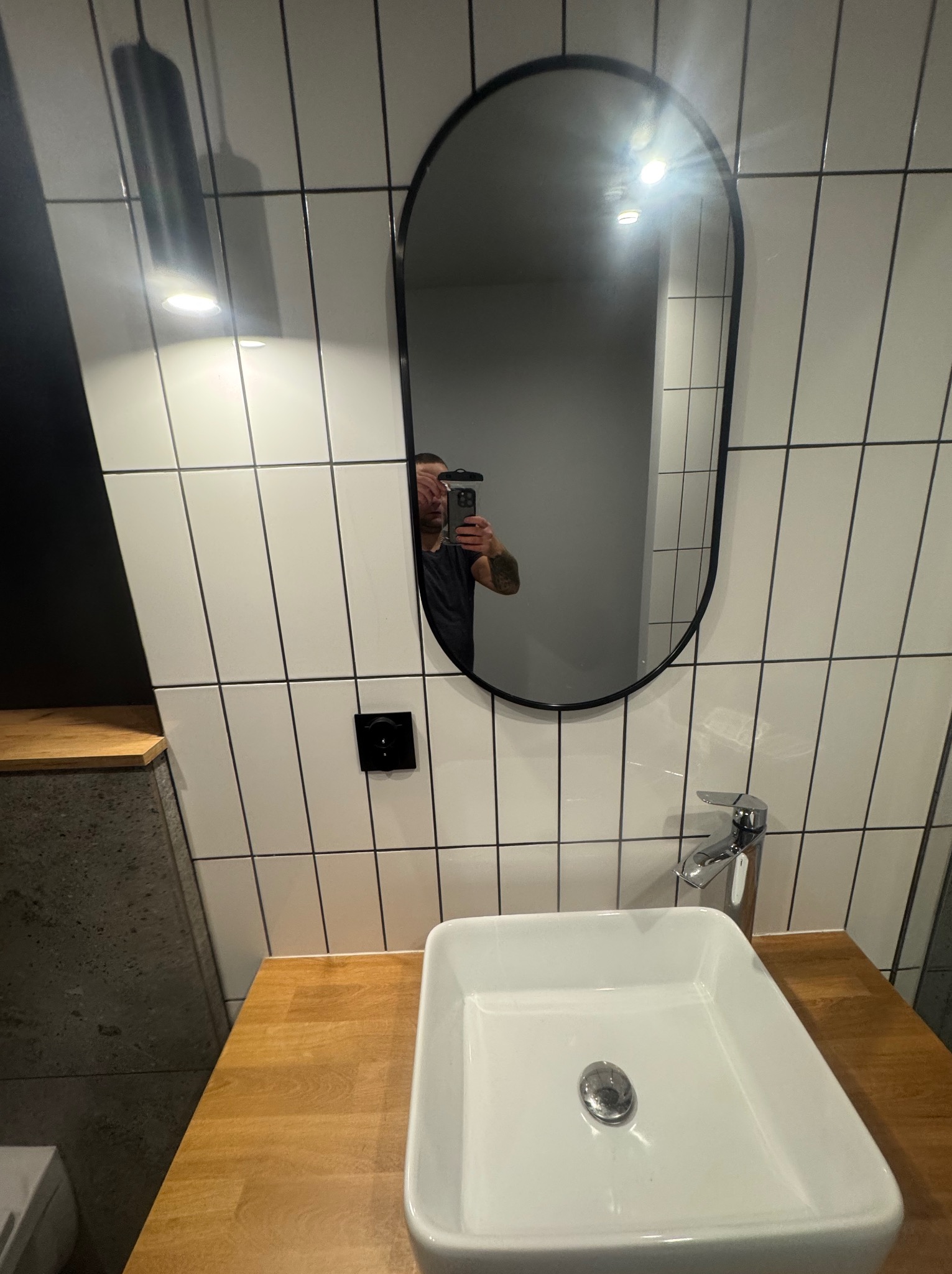 Nowoczesna łazienka z owalnym lustrem w czarnej ramie, białą umywalką na drewnianym blacie i białymi płytkami z czarną fugą. W lustrze odbija się osoba robiąca zdjęcie.