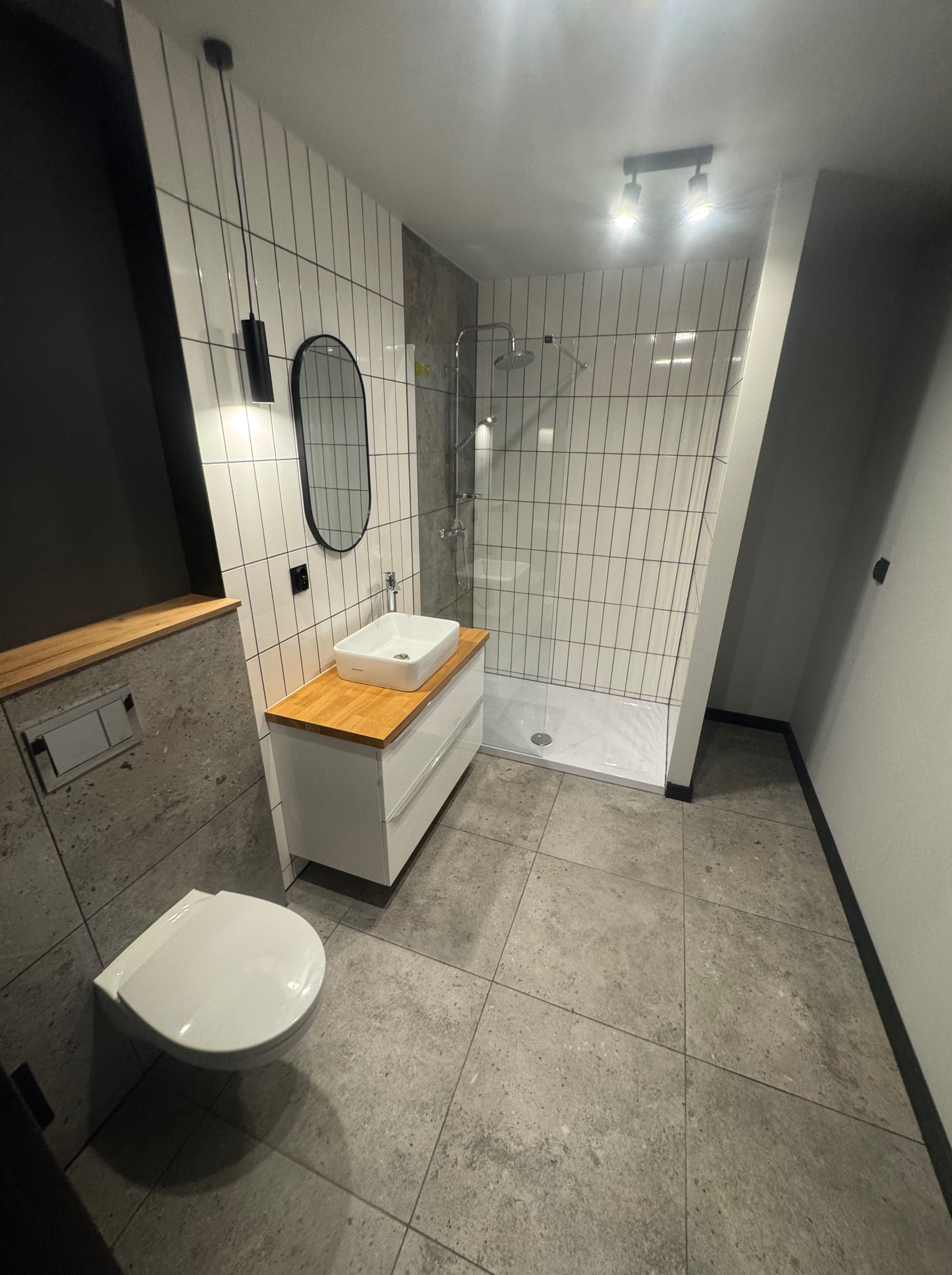 Nowoczesna łazienka z kabiną prysznicową typu walk-in, białą umywalką na szafce z drewnianym blatem i wiszącą toaletą. Dominuje szaro-biała kolorystyka z czarnymi akcentami.