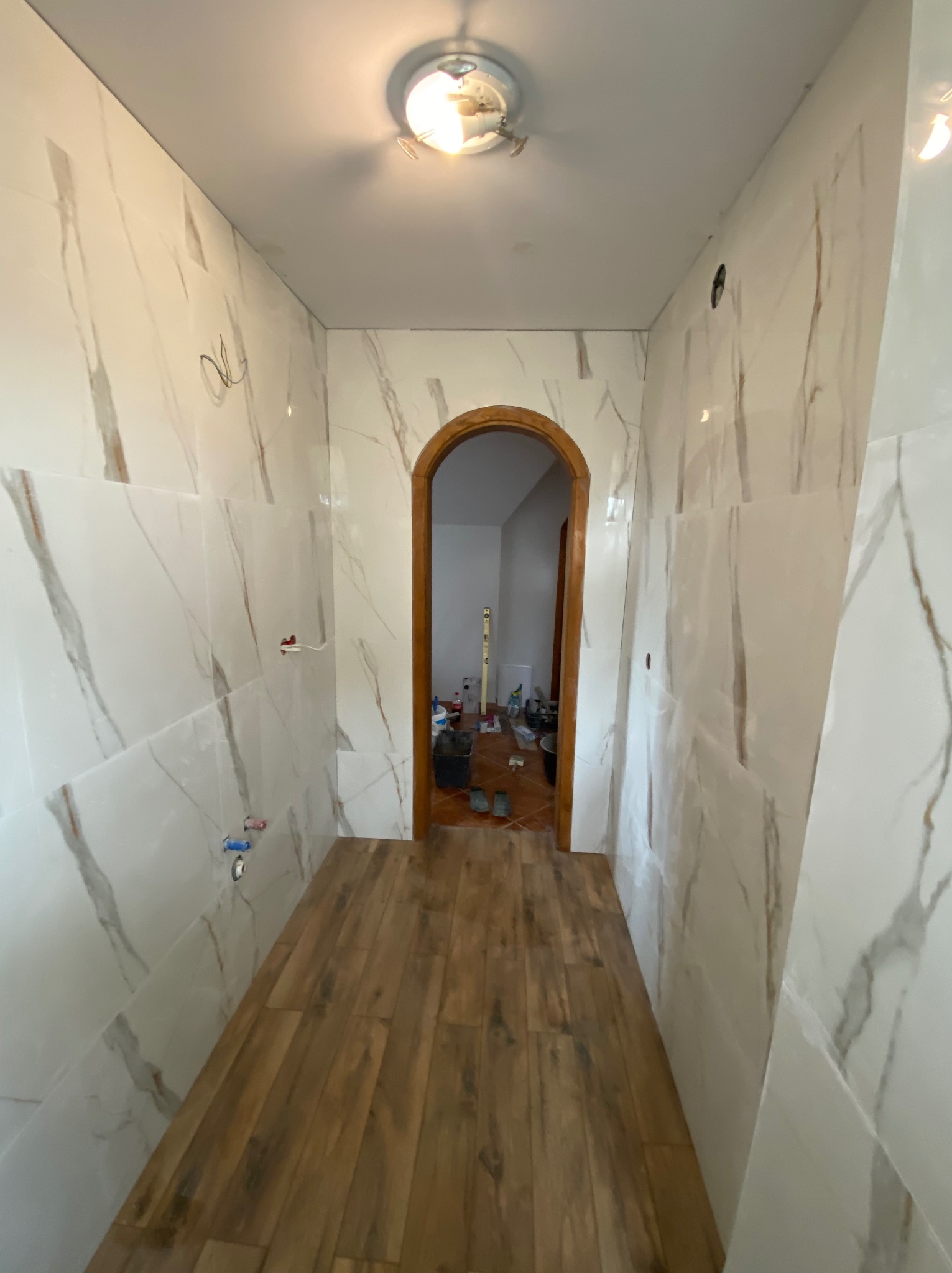 Wnętrze łazienki w trakcie remontu: marmurowe płytki na ścianach, drewnopodobne panele podłogowe, widoczne instalacje i narzędzia w przejściu.