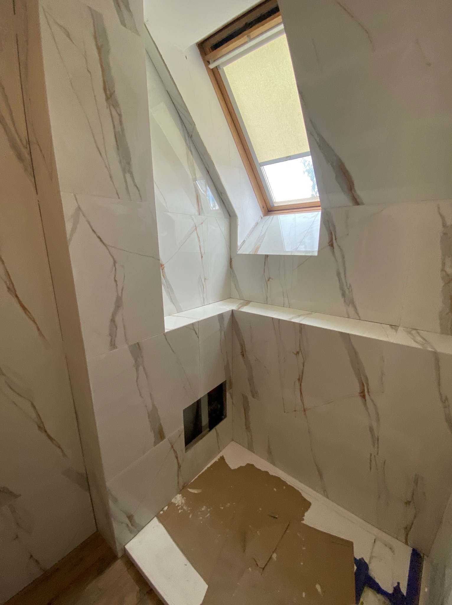 Ukończona łazienka na poddaszu z oknem dachowym. Białe płytki imitujące marmur z złotymi żyłami na ścianach i częściowo na podłodze. Fragment zabezpieczonej podłogi.