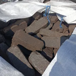 Brykiet drzewny w worku jutowym, widoczne niebieskie sznurki, oświetlenie słoneczne.