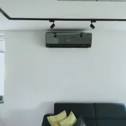 Nowoczesny czarny klimatyzator zamontowany na białej ścianie nad ciemną sofą z żółtymi poduszkami, oświetlony reflektorami na szynie.