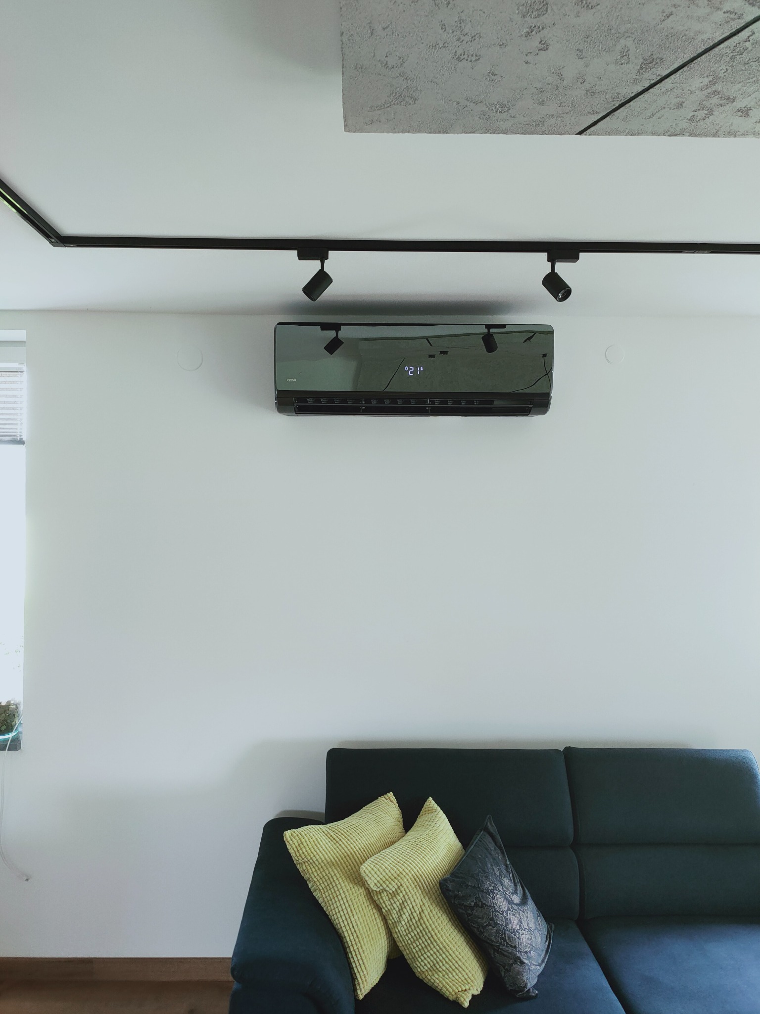 Nowoczesny czarny klimatyzator zamontowany na białej ścianie nad ciemną sofą z żółtymi poduszkami, oświetlony reflektorami na szynie.