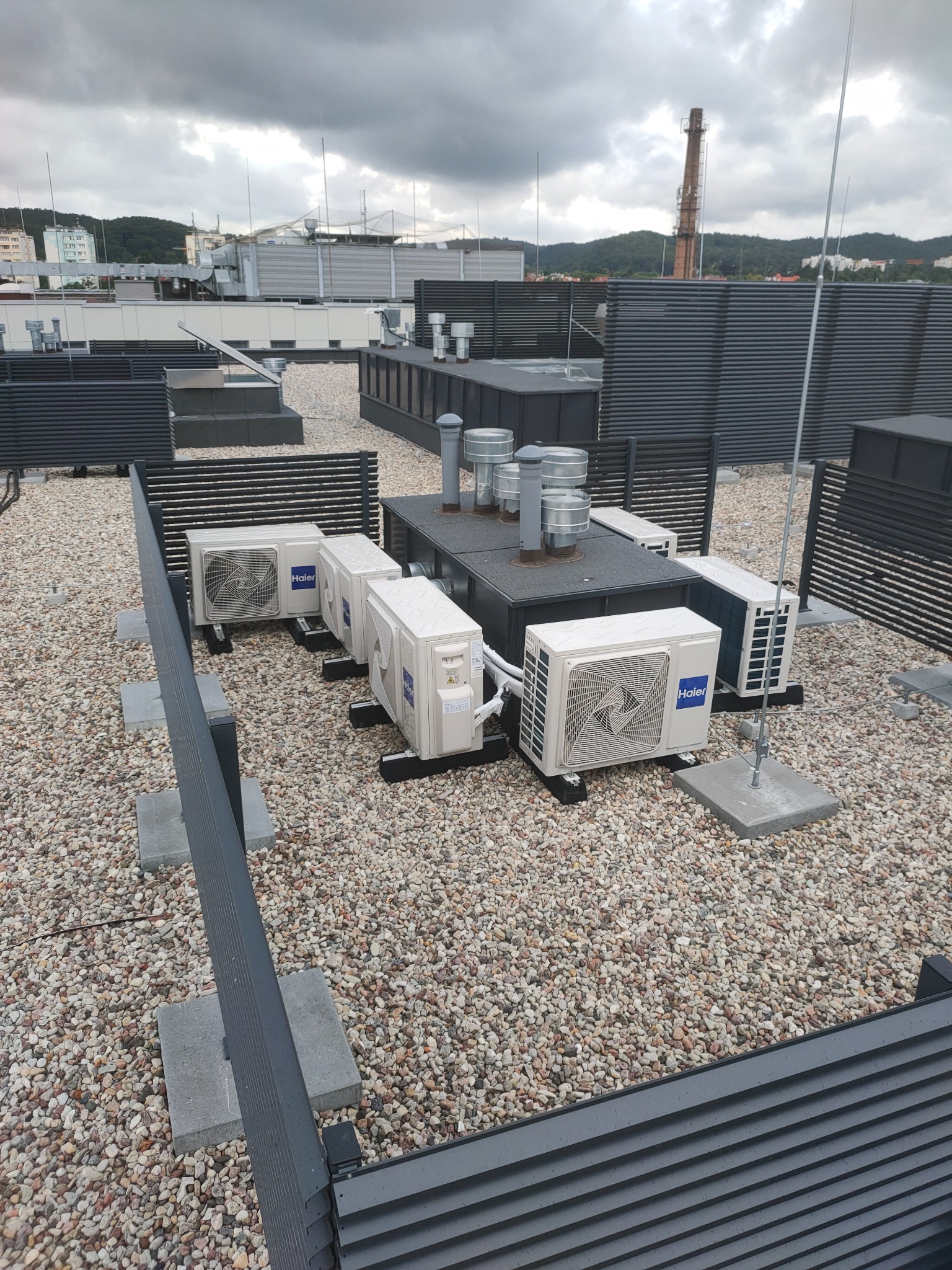 Rzut z góry na instalację klimatyzacji marki Haier na dachu budynku pokrytym żwirem, z widocznymi elementami wentylacyjnymi i kominem w tle, pod pochmurnym niebem.