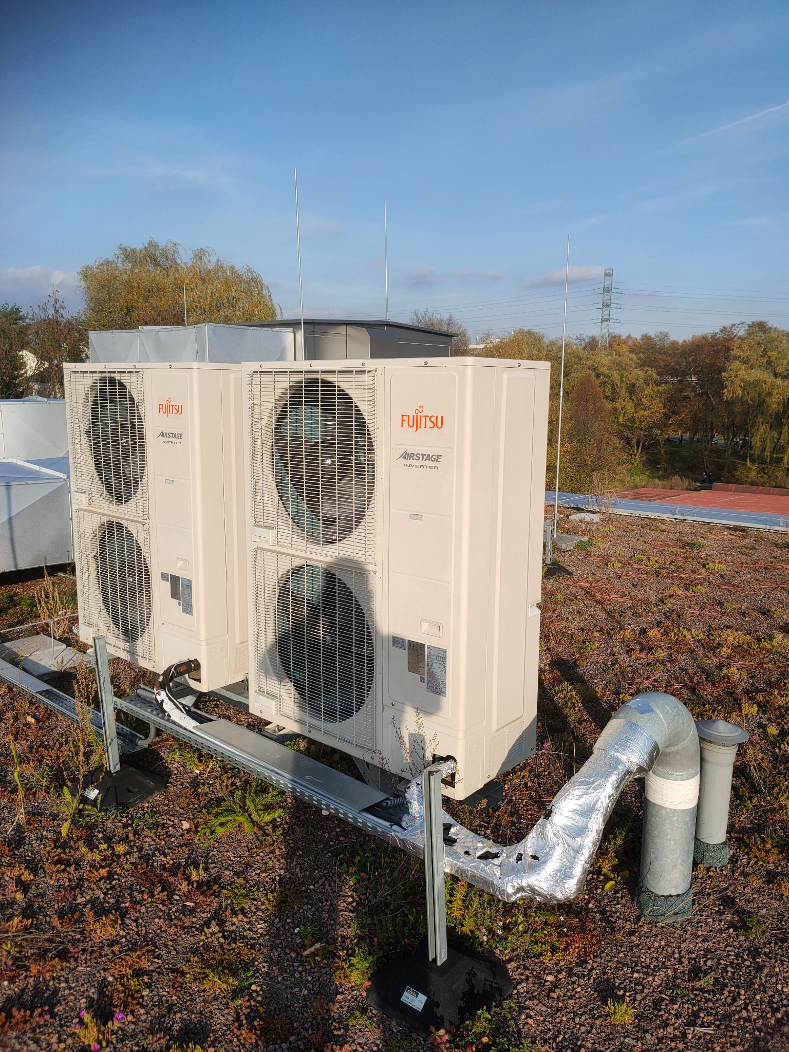 Dwa agregaty klimatyzacyjne Fujitsu Airstage Inverter zamontowane na metalowej konstrukcji na zielonym dachu z widocznymi przewodami i elementami instalacji, w tle drzewa i słup wysokiego napięcia.