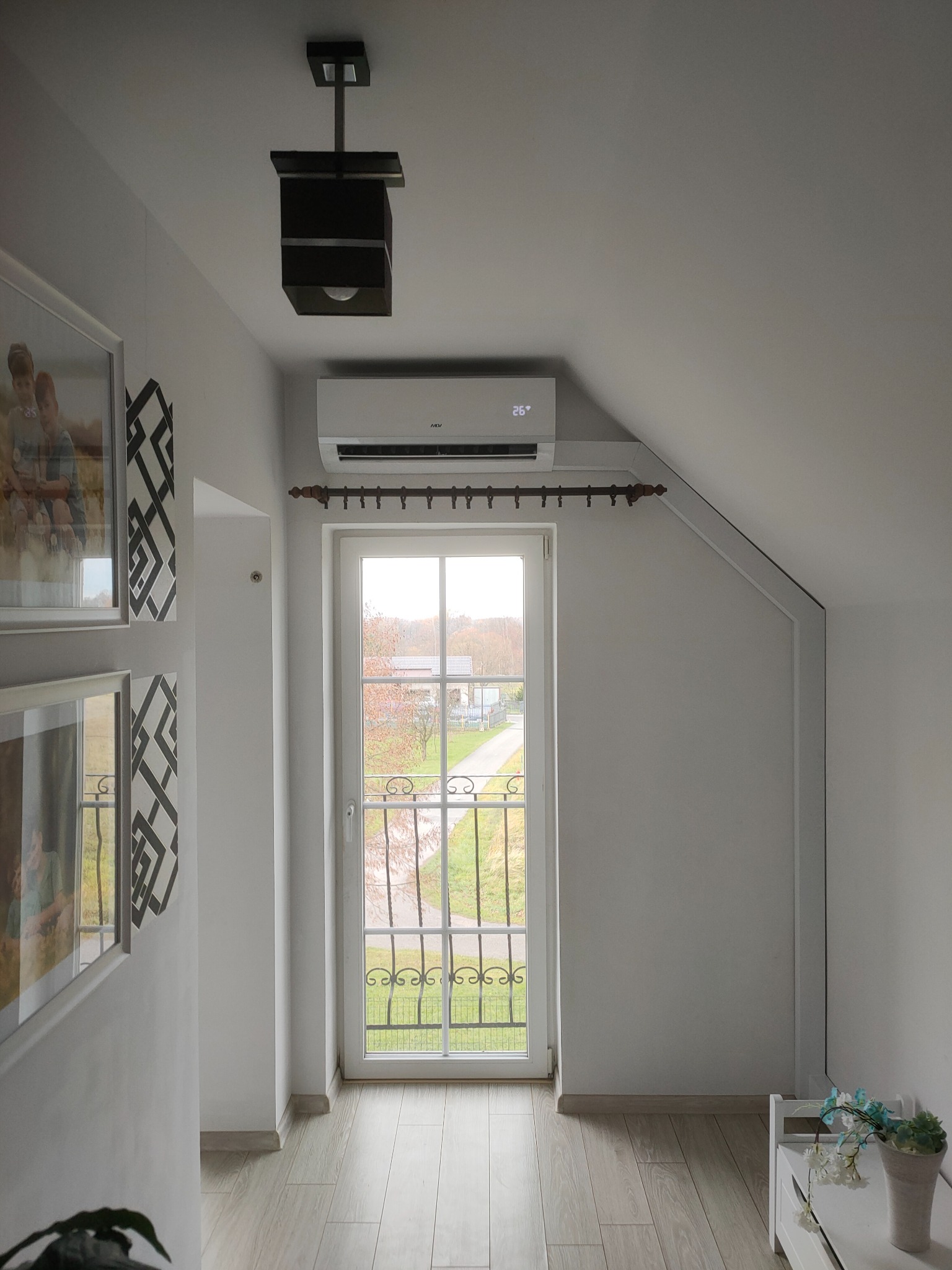 Wnętrze poddasza z zainstalowanym klimatyzatorem nad oknem, widoczny termometr na urządzeniu wskazujący temperaturę 26 stopni Celsjusza, drewniana podłoga, dekoracje ścienne i lampa sufitowa.