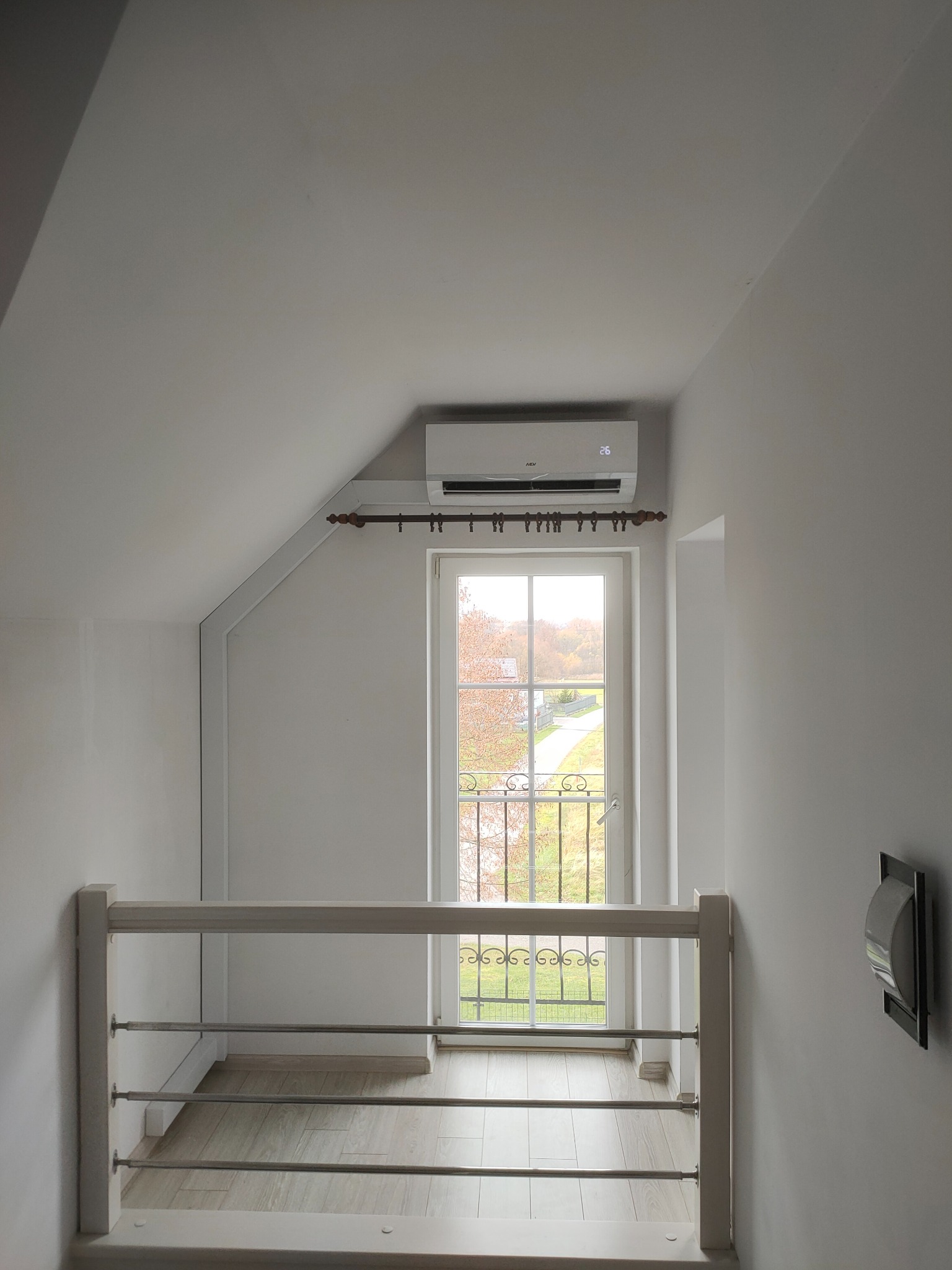 Biały klimatyzator na poddaszu zamontowany nad oknem balkonowym z ozdobną balustradą, widok z góry na drewnianą podłogę.