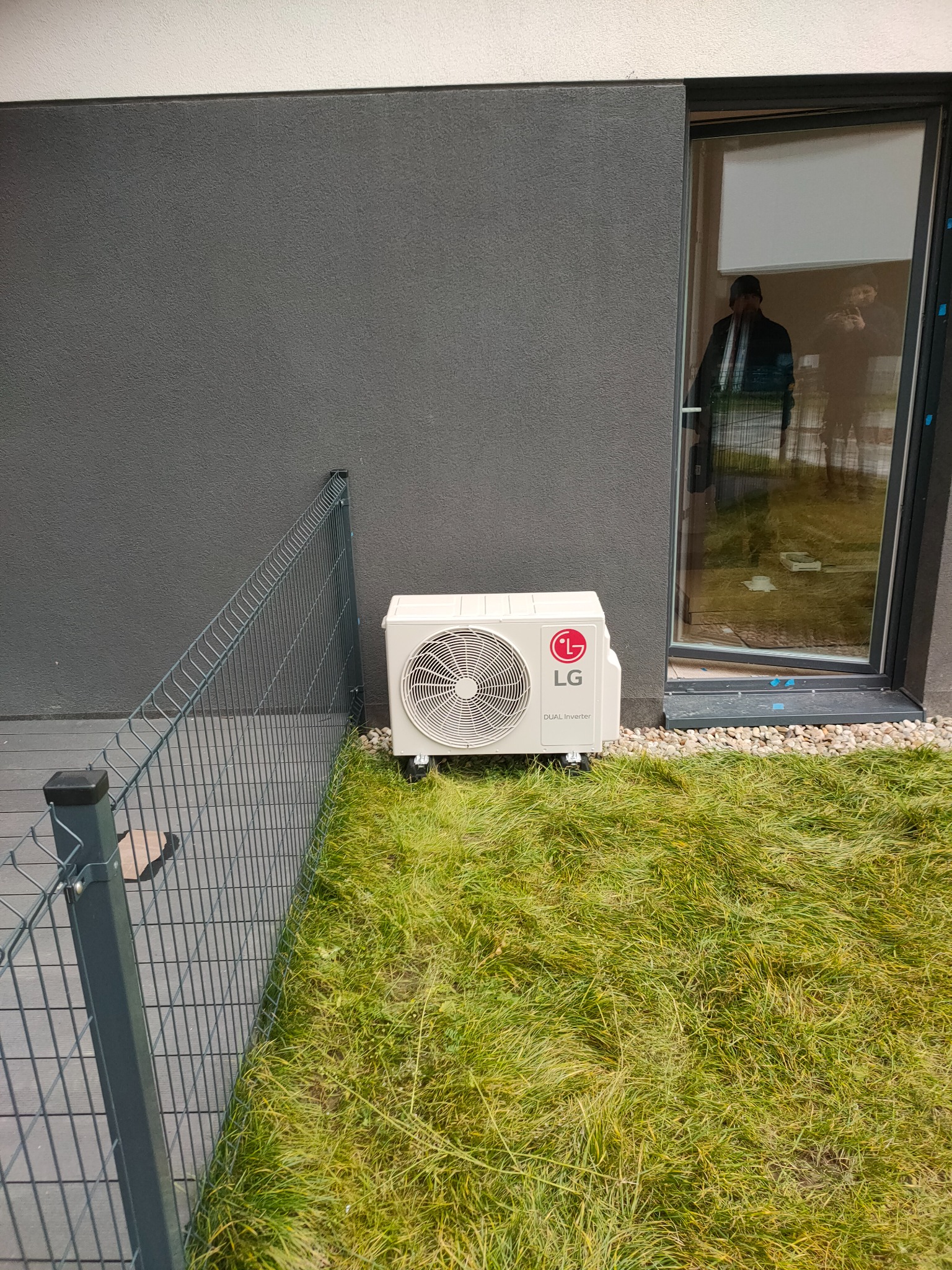 Zewnętrzna jednostka klimatyzacji LG Dual Inverter umieszczona na trawie obok szarej ściany budynku i okna z odbijającą się sylwetką osoby.