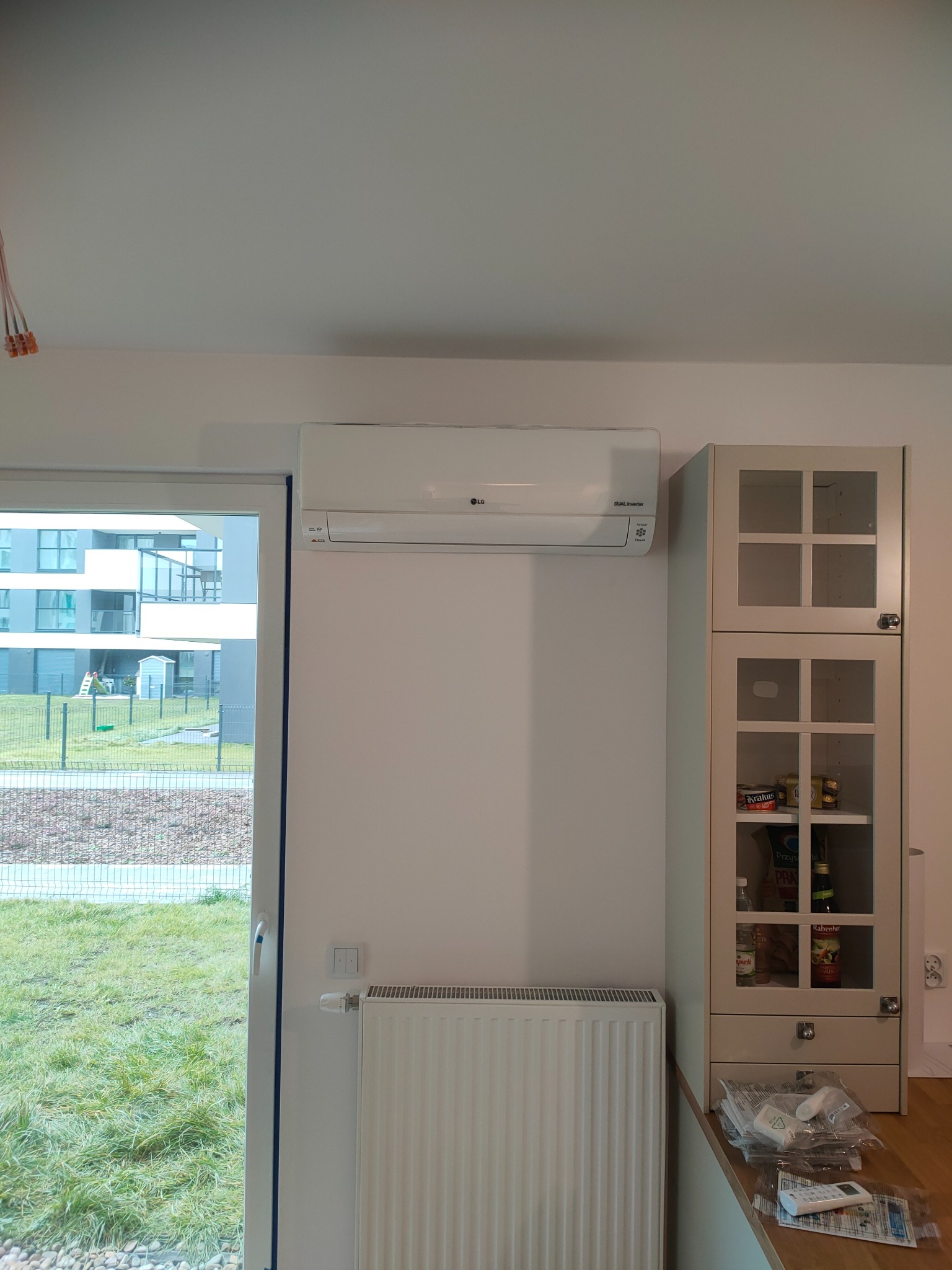 Biały klimatyzator LG zamontowany na ścianie w jasnym pomieszczeniu z widokiem na nowoczesny budynek przez okno, obok szafka z przeszklonymi drzwiczkami i grzejnik.