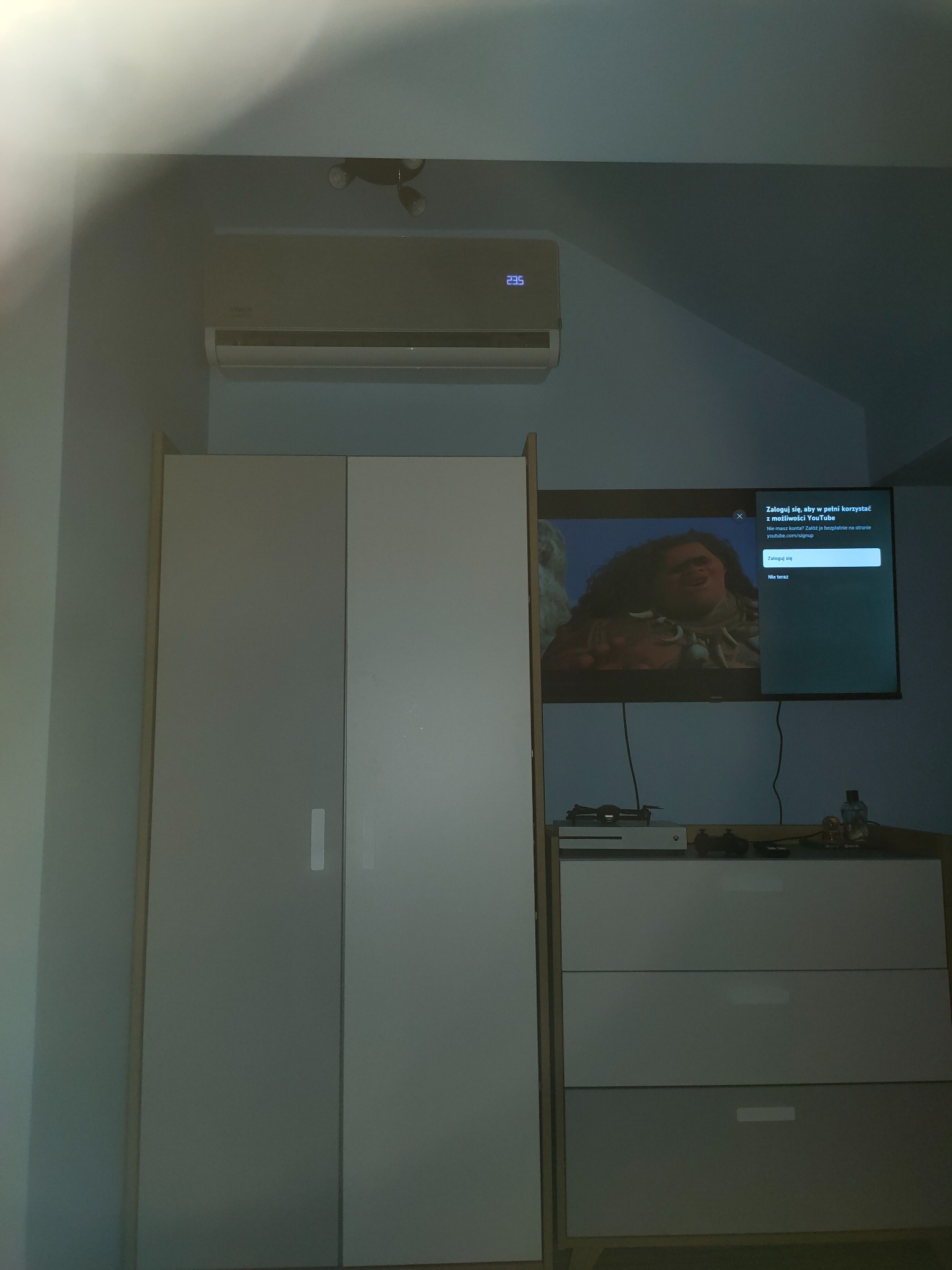 Szare urządzenie klimatyzacyjne zamontowane na ścianie w pokoju, widoczne nad szafą i komodą z telewizorem wyświetlającym film animowany.