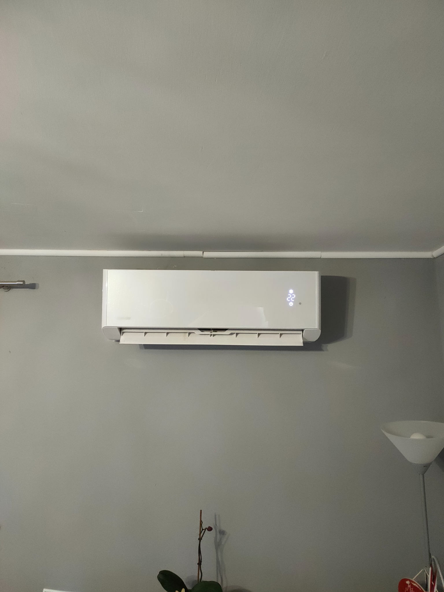 Biały klimatyzator ścienny zamontowany w szarym pokoju z widocznym wyświetlaczem temperatury, lampą stojącą i rośliną doniczkową w tle.