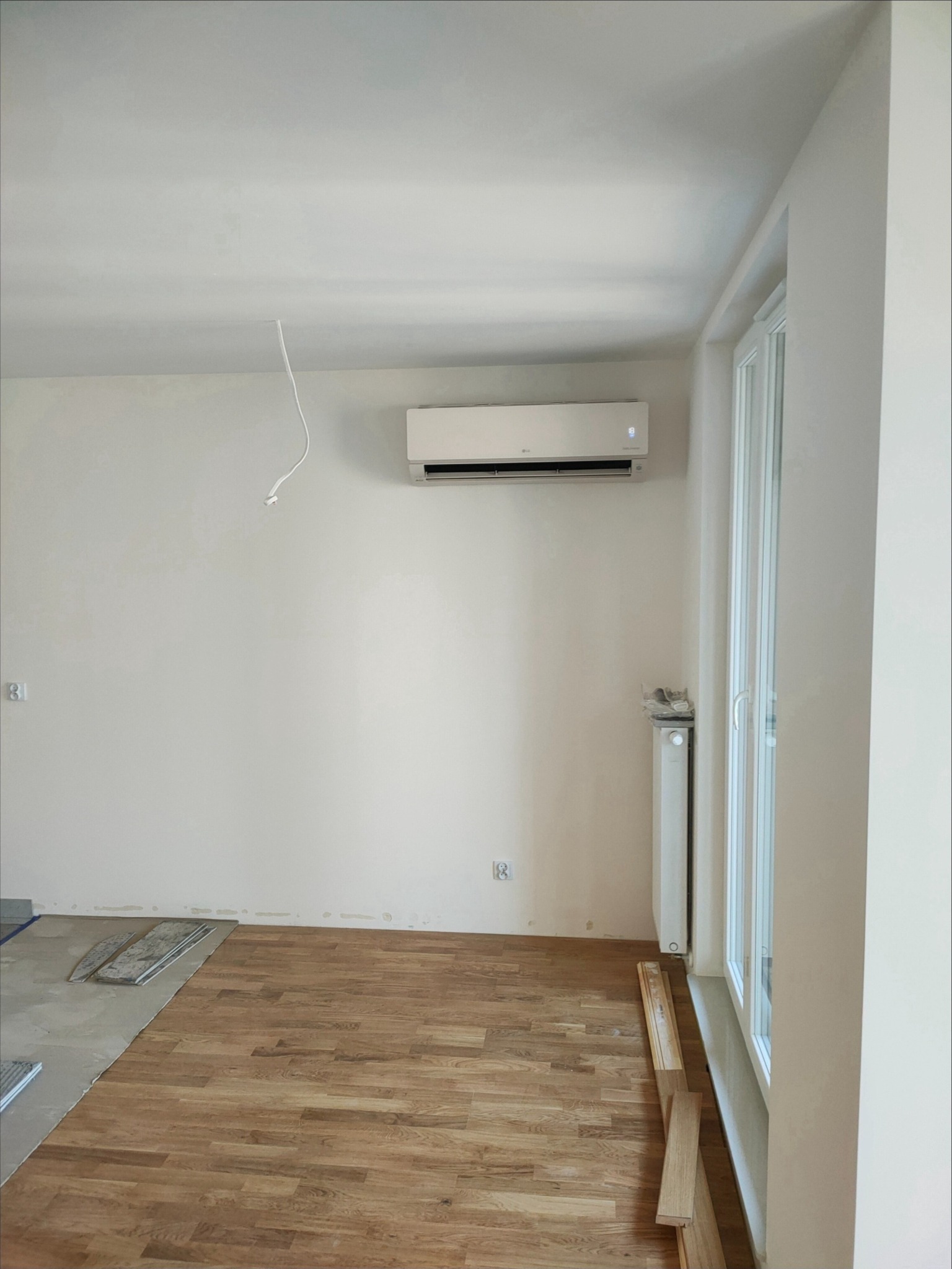 Wnętrze pokoju w trakcie remontu z zamontowanym klimatyzatorem LG na ścianie, widoczne niedokończone podłogi, grzejnik i kabel zwisający z sufitu.