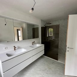 BN-INSTAL - Nowoczesna łazienka w trakcie wykończenia, z podwójną białą umywalką nablatową z złotą armaturą, dużym lustrem z oświetleniem, szklaną kabiną prysznicową i marmurową posadzką.