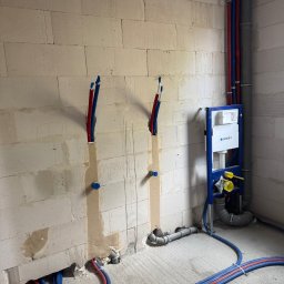 BN-INSTAL - Surowa ściana z betonu komórkowego z instalacją hydrauliczną w kolorze niebieskim i czerwonym, widoczny stelaż podtynkowy Geberit oraz rury kanalizacyjne.