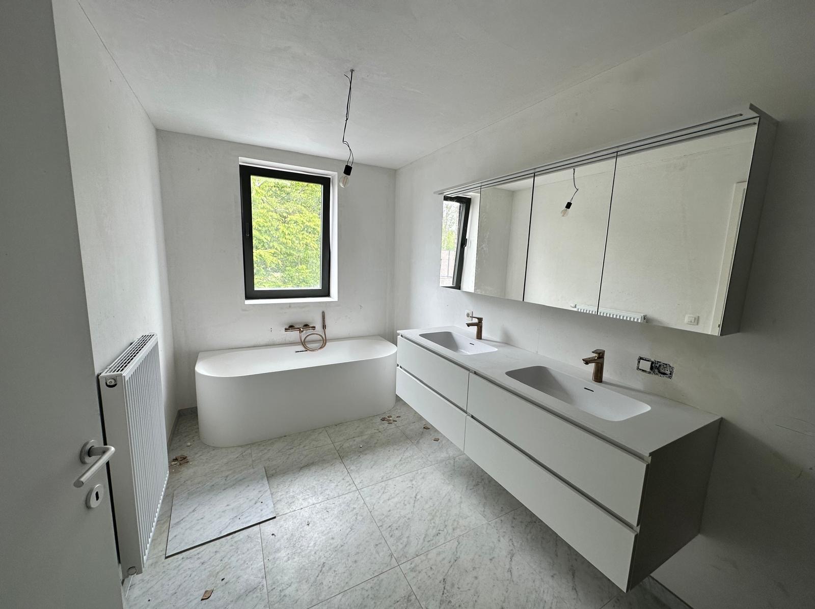 Łazienka w trakcie remontu: biała wanna wolnostojąca pod oknem z widokiem na zieleń, podwójna umywalka z szafką i lustrem, marmurowe płytki podłogowe, grzejnik na ścianie, widoczne niedociągnięcia...