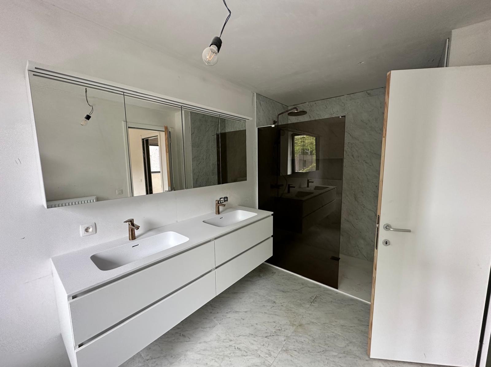 Nowoczesna łazienka w trakcie wykończenia, z podwójną białą umywalką nablatową z złotą armaturą, dużym lustrem z oświetleniem, szklaną kabiną prysznicową i marmurową posadzką.