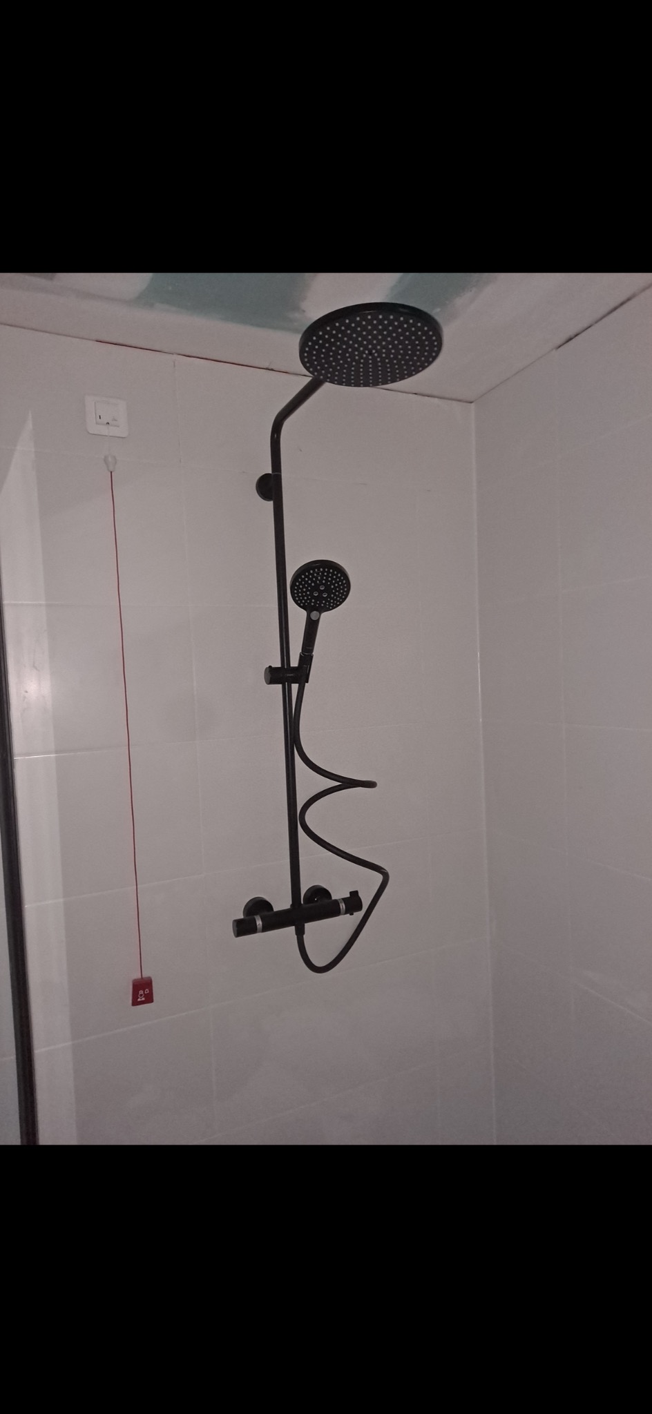 Czarny zestaw prysznicowy z deszczownicą i słuchawką, zamontowany na białej, kafelkowanej ścianie w łazience w trakcie remontu, widoczne niedokończone łączenia płyt na suficie.