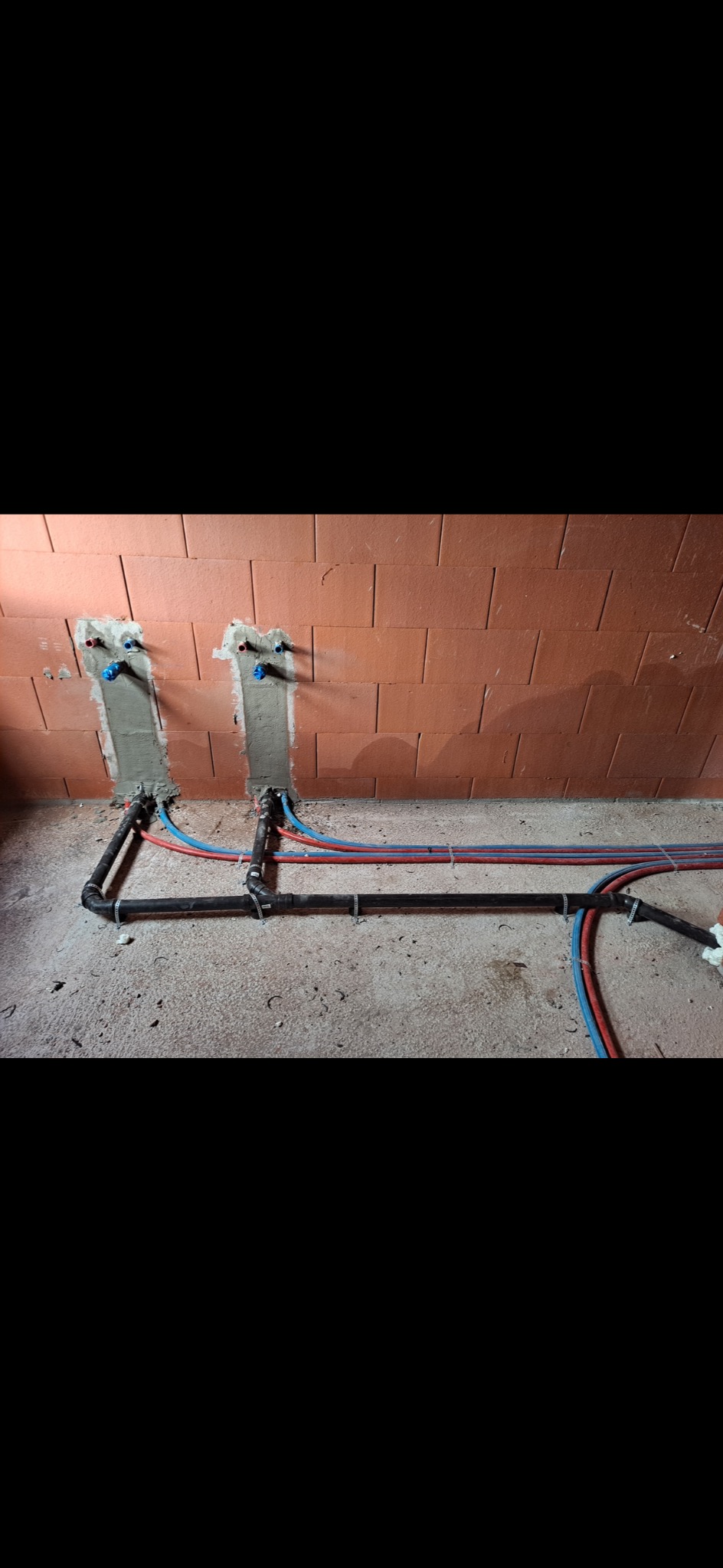 Surowa ściana z cegły z wmurowanymi rurami wodnymi i grzewczymi, widoczne czarne rury kanalizacyjne na podłodze w trakcie montażu.