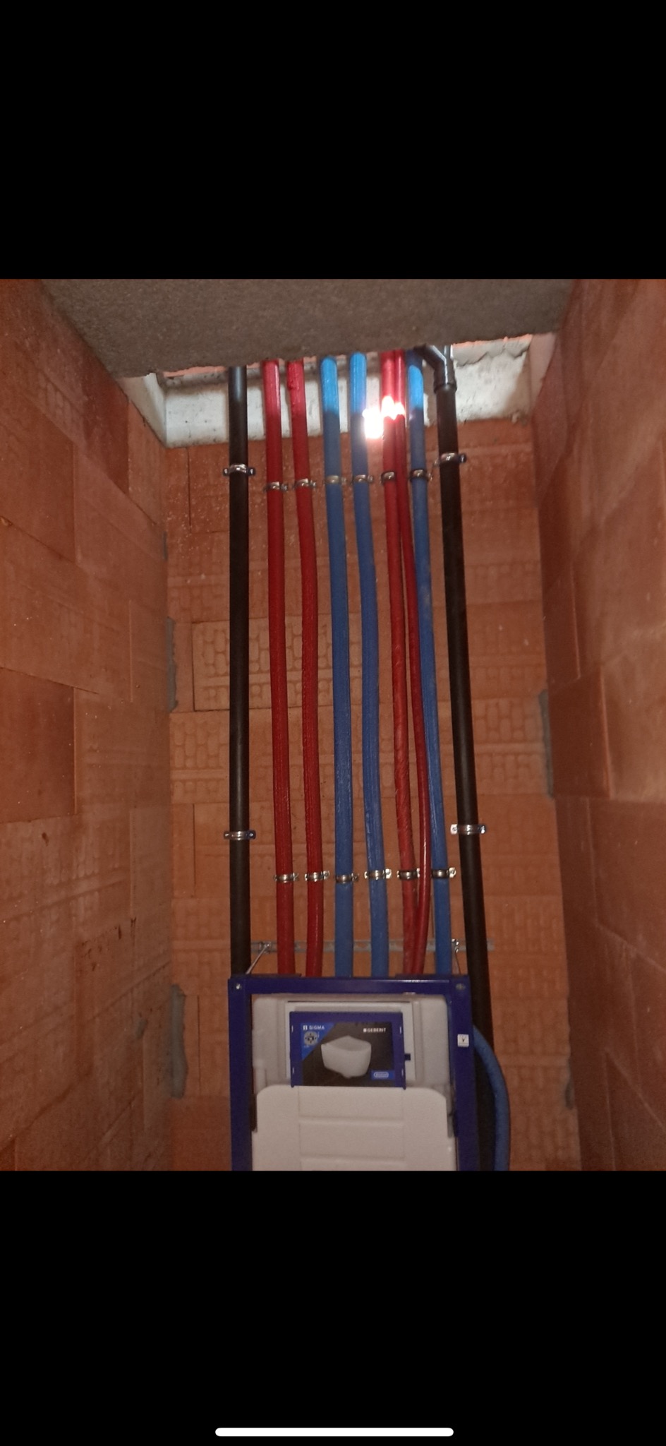 Instalacja podtynkowa WC Geberit Sigma z doprowadzonymi rurami PEX w kolorach czerwonym i niebieskim, przymocowanymi do ściany z cegły dziurawki metalowymi uchwytami.
