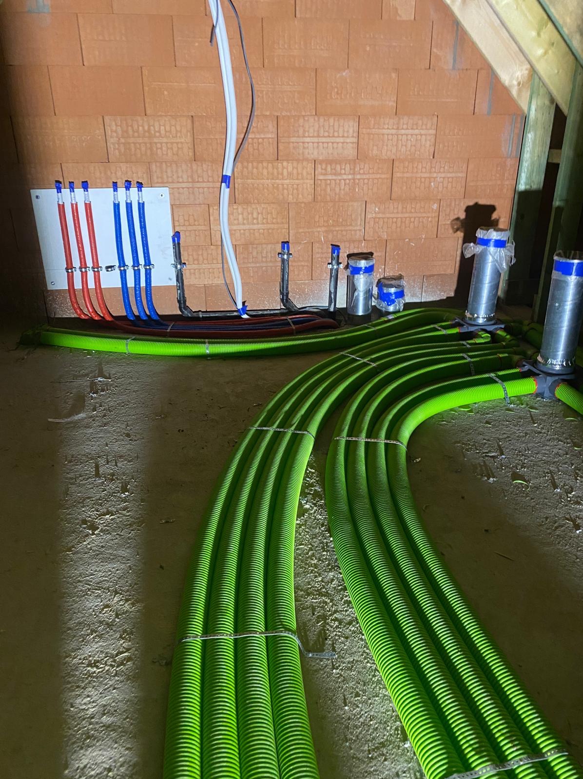 Rozprowadzenie zielonych rur ogrzewania podłogowego w surowym stanie, widoczne kolektory i instalacja hydrauliczna na ścianie z cegły.