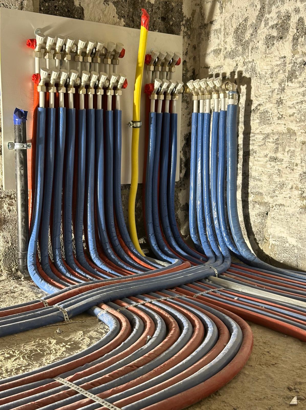 Rozdzielacz instalacji grzewczej z licznymi rurami w izolacji koloru niebieskiego i czerwonego, ułożonymi równolegle na podłodze w surowym pomieszczeniu z kamiennymi ścianami.