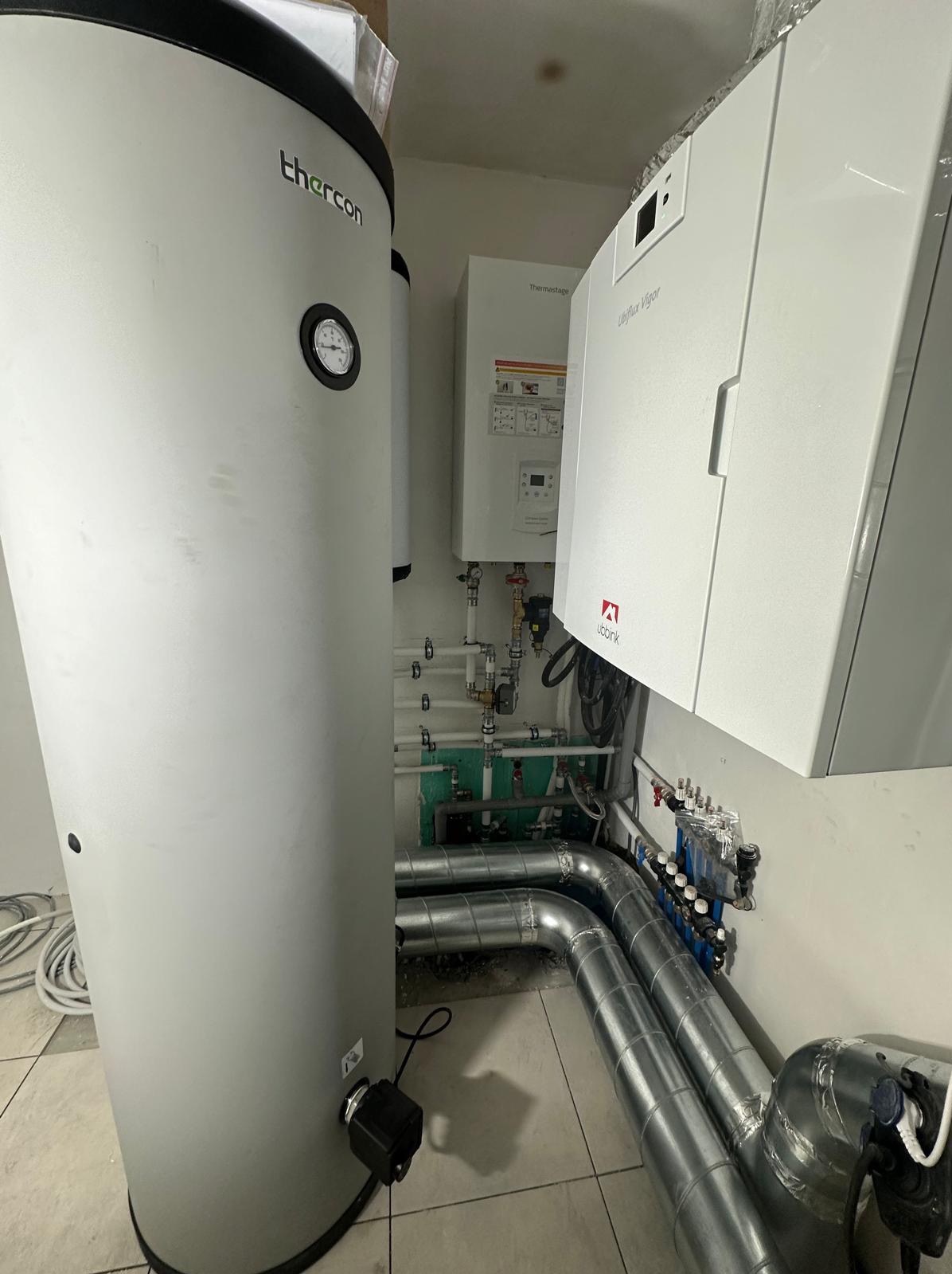 Szary, cylindryczny zbiornik Thermancon z manometrem obok białego, wiszącego pieca Uniflux Vigor z widocznymi rurami instalacji grzewczej i rozdzielaczem w pomieszczeniu technicznym.