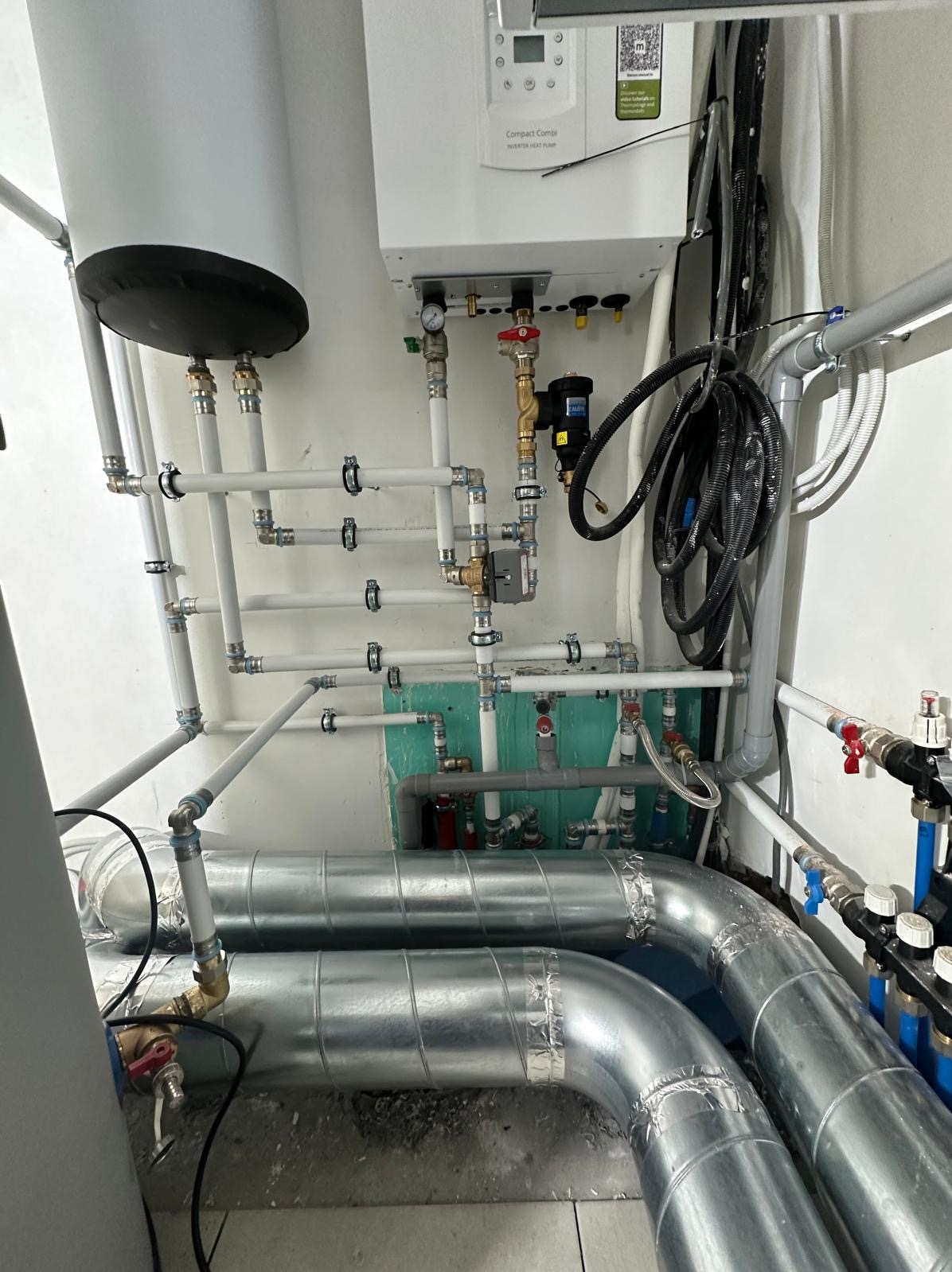 Kotłownia z zainstalowanym piecem i rozbudowaną instalacją grzewczą, widoczne białe rury z licznymi złączkami, izolowane rury metalowe, oraz elementy sterujące. Na ścianie zamontowany piec gazowy...