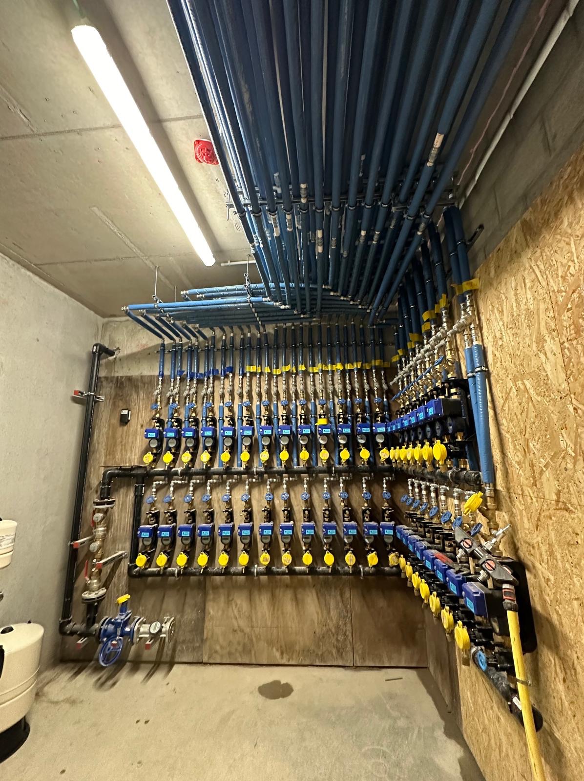 Rozbudowana instalacja hydrauliczna z licznymi niebieskimi rurami i zaworami z żółtymi pokrętłami, zamontowana na ścianie z płyty OSB w surowym pomieszczeniu z betonowym sufitem.