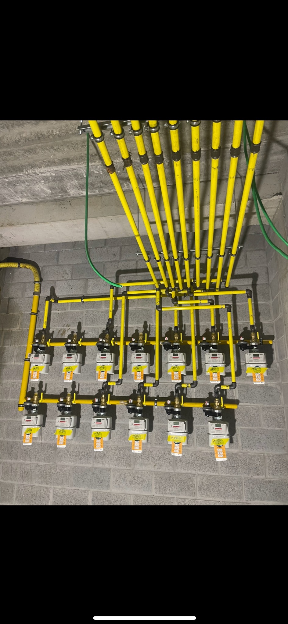 Rząd dziesięciu nowych, zamontowanych gazomierzy z żółtymi rurami gazowymi, przymocowanych do szarej ściany z cegieł w piwnicy.
