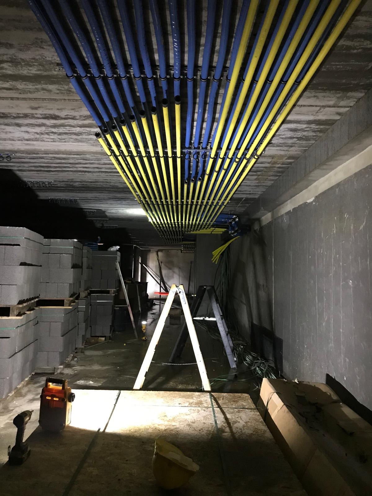 Instalacja rzędów niebieskich i żółtych rur pod betonowym sufitem, widoczne drabiny, narzędzia i materiały budowlane na podłodze.