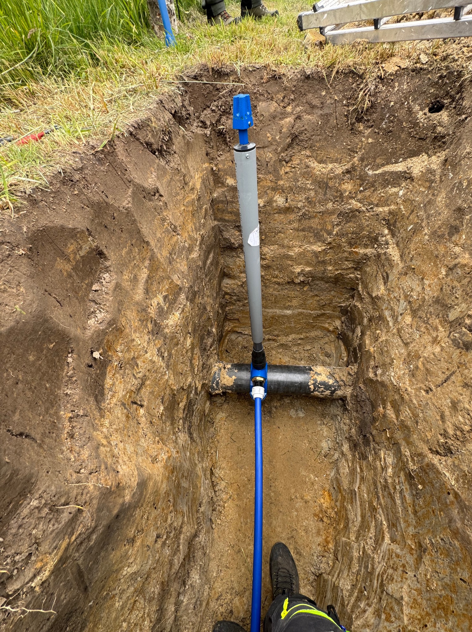 Instalacja rur w wykopie ziemnym, widoczne połączenia i fragment instalacji nad ziemią. Detal: buty montera w rogu kadru.