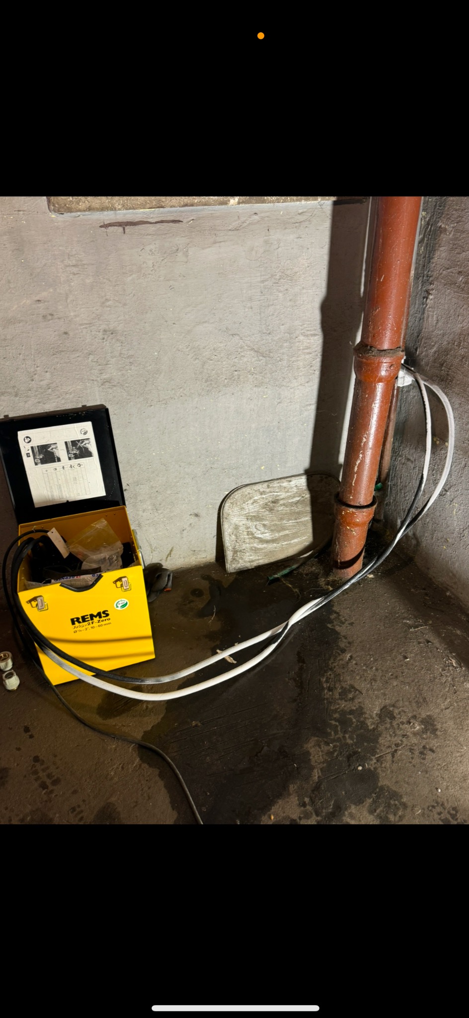 Brązowa rura kanalizacyjna w rogu pomieszczenia, obok żółta skrzynka narzędziowa REMS z instrukcją i przewodami. Podłoże zabrudzone, ściany betonowe.