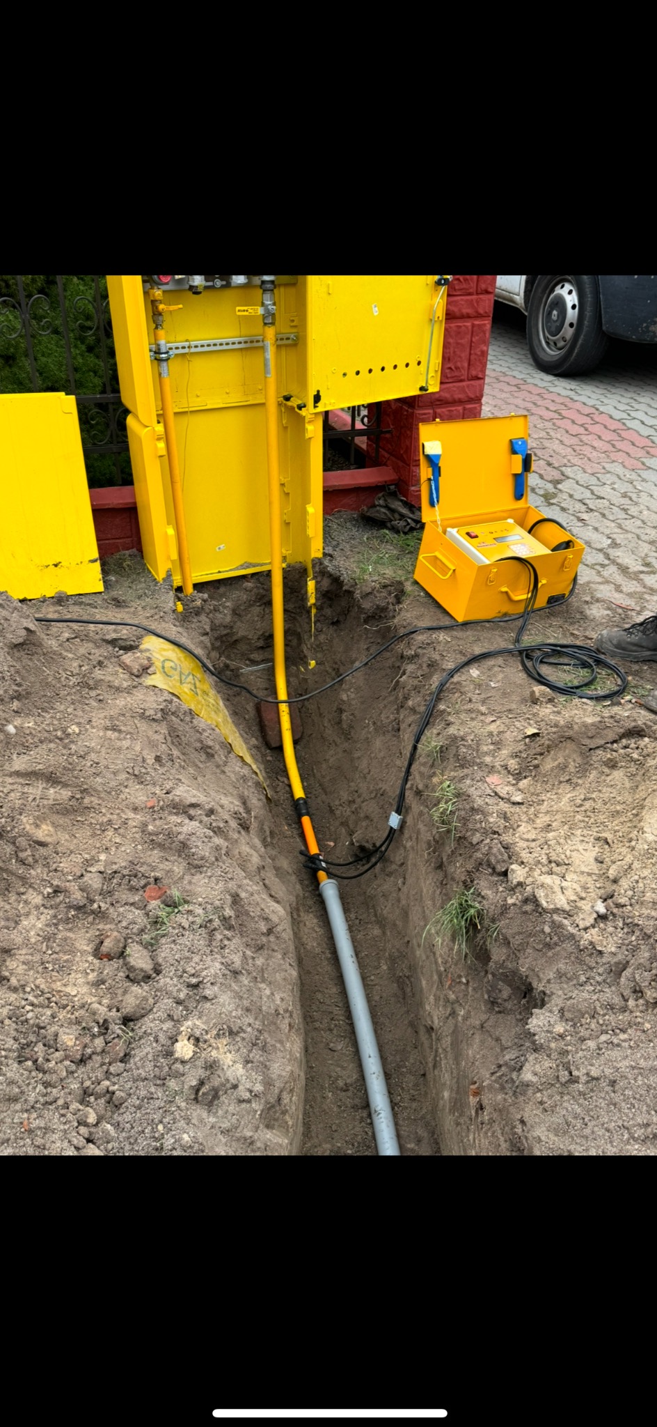 Wykop z ułożoną szarą rurą i żółtym kablem. Obok żółta skrzynka gazowa i narzędzia pomiarowe. Prace ziemne przyłącza gazowego w trakcie realizacji.