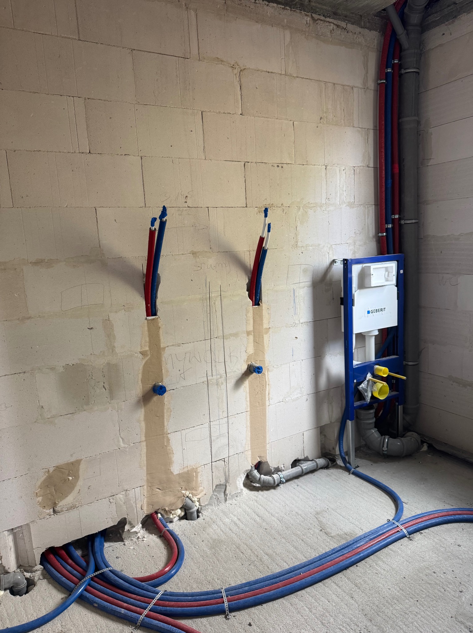 Surowa ściana z betonu komórkowego z instalacją hydrauliczną w kolorze niebieskim i czerwonym, widoczny stelaż podtynkowy Geberit oraz rury kanalizacyjne.