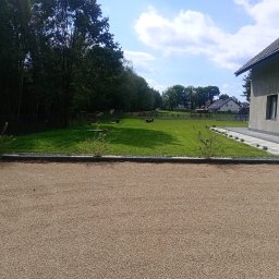 C&W Building sp. z o.o. - Nowoczesny dom z zadbanym trawnikiem, utwardzonym podjazdem z kamienia i minimalistyczną architekturą ogrodu. Widok na zielony teren z drzewami w tle.