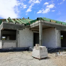 Stan surowy otwarty domu jednorodzinnego w Gogołowej. Widoczne ściany z betonu kom&oacute;rkowego, więźba dachowa zaimpregnowana na zielono i paleta z bloczkami.