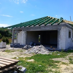 C&W Building sp. z o.o. - Stan surowy otwarty domu z białego bloczka, z zielonymi elementami więźby dachowej, na tle błękitnego nieba. Widoczne materiały budowlane i otoczenie w trawie.