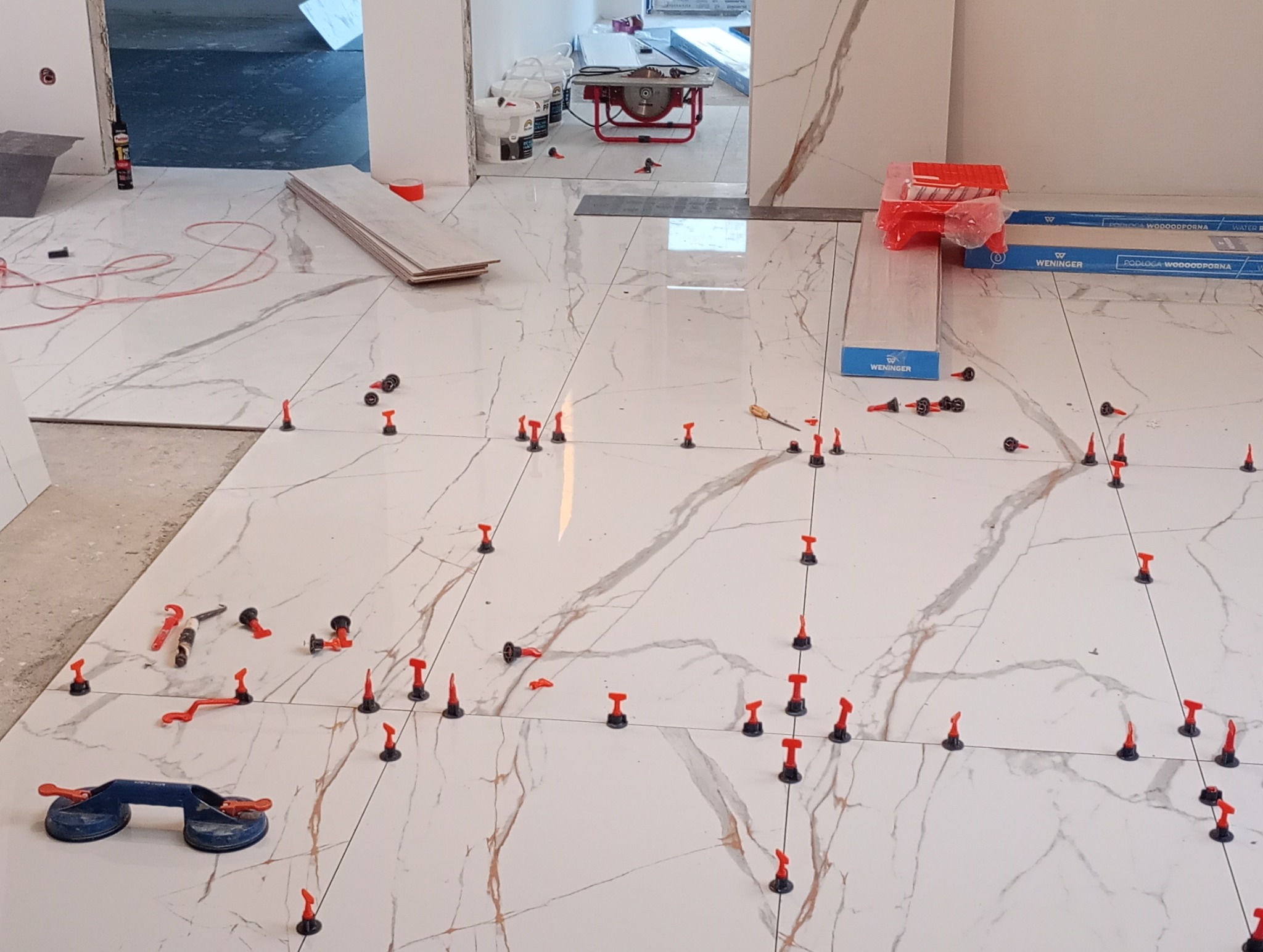 Układanie płytek podłogowych z marmurowym wzorem, widoczne narzędzia i system poziomowania płytek. Częściowo ułożona podłoga w jasnym pomieszczeniu.