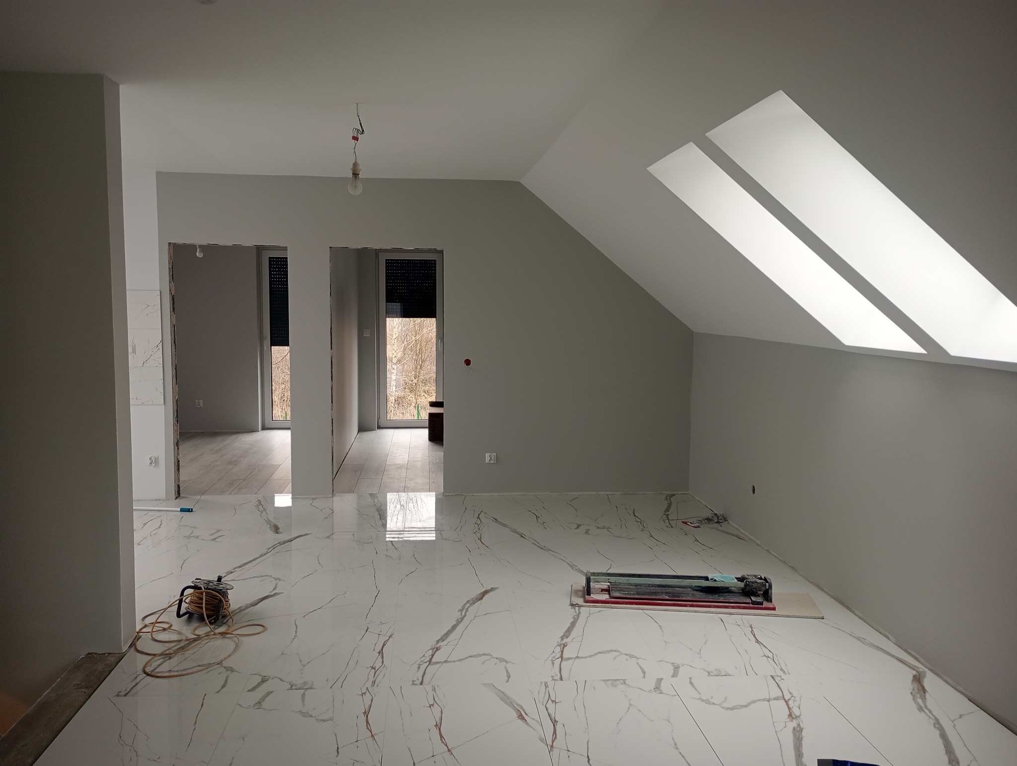 Jasne, wykończone poddasze z marmurową podłogą i oknami dachowymi. Widoczny sprzęt do cięcia płytek i zwinięty kabel. W tle otwarte drzwi do innych pomieszczeń.