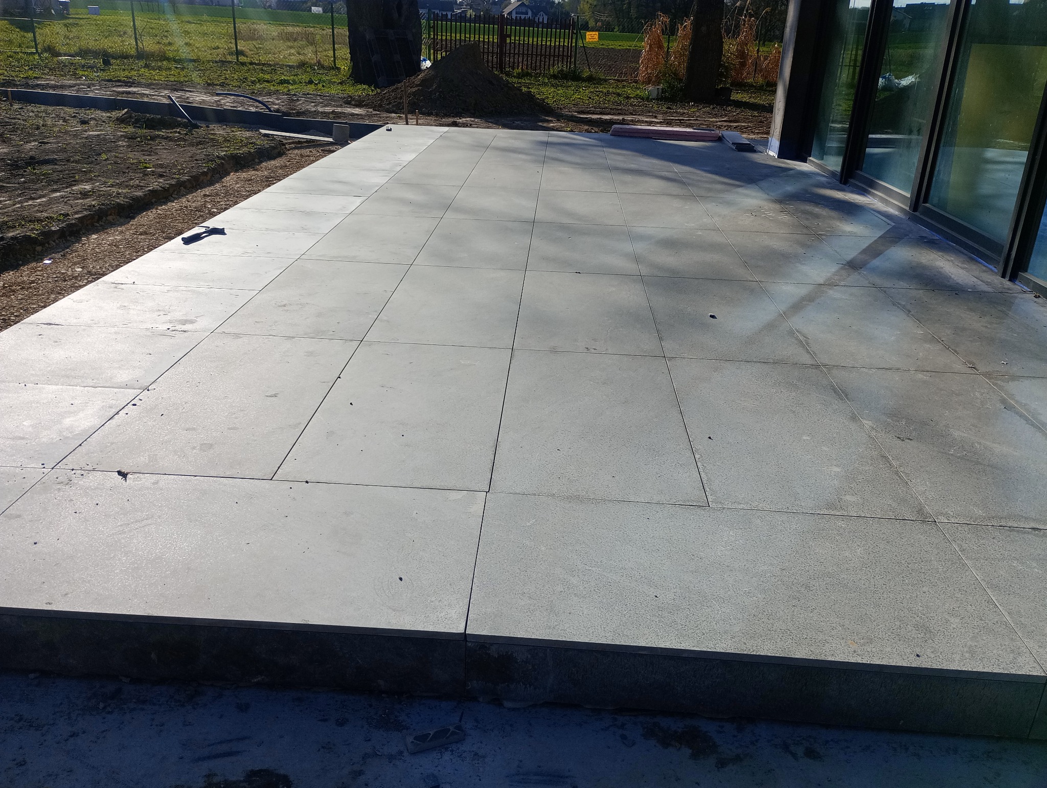 Ułożona, szara, kwadratowa nawierzchnia tarasu z płytek, widok z góry. W tle trawa, ziemia i budynek z dużymi oknami. Na pierwszym planie krawędź tarasu.