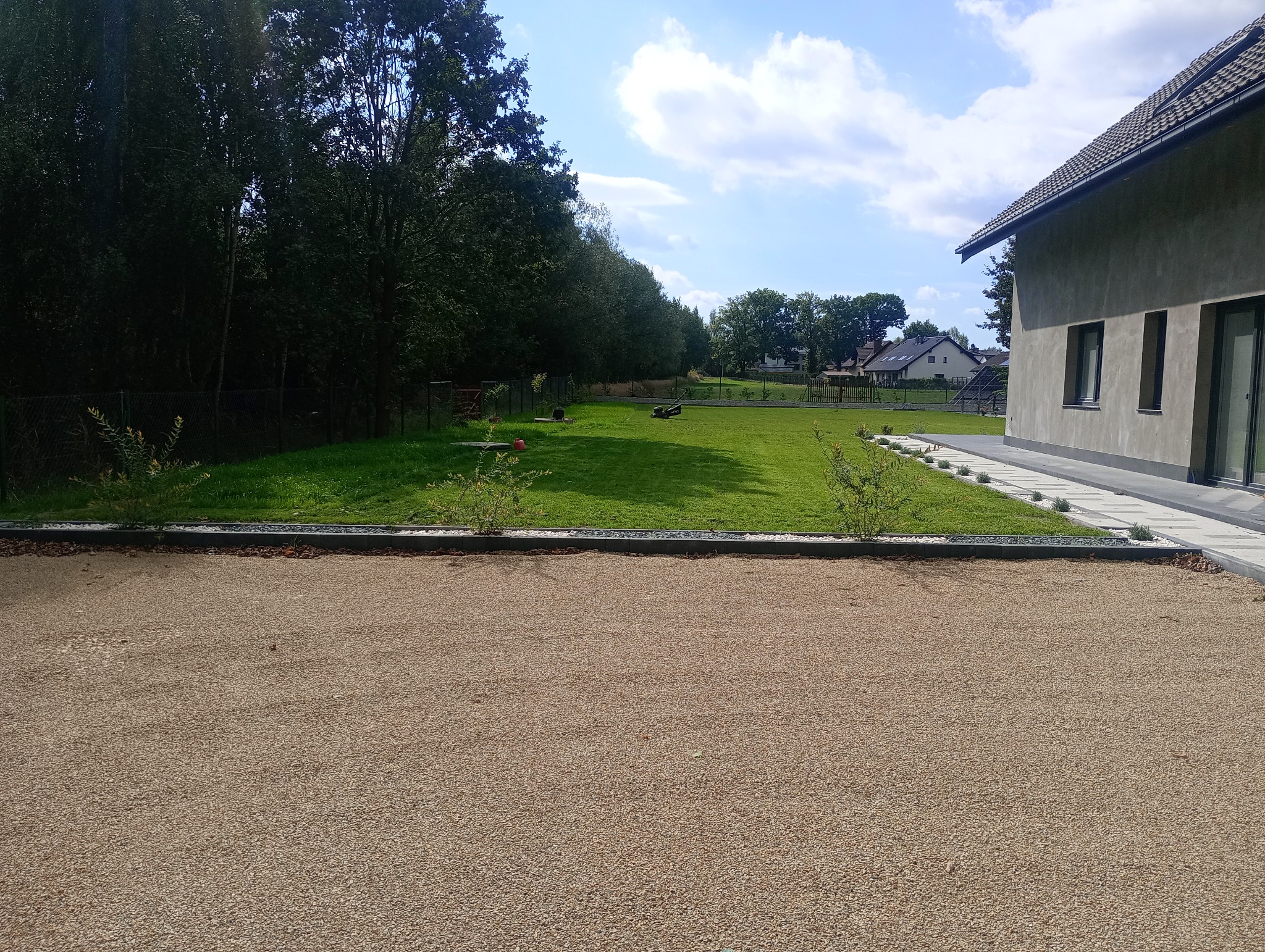 Nowoczesny dom z zadbanym trawnikiem, utwardzonym podjazdem z kamienia i minimalistyczną architekturą ogrodu. Widok na zielony teren z drzewami w tle.