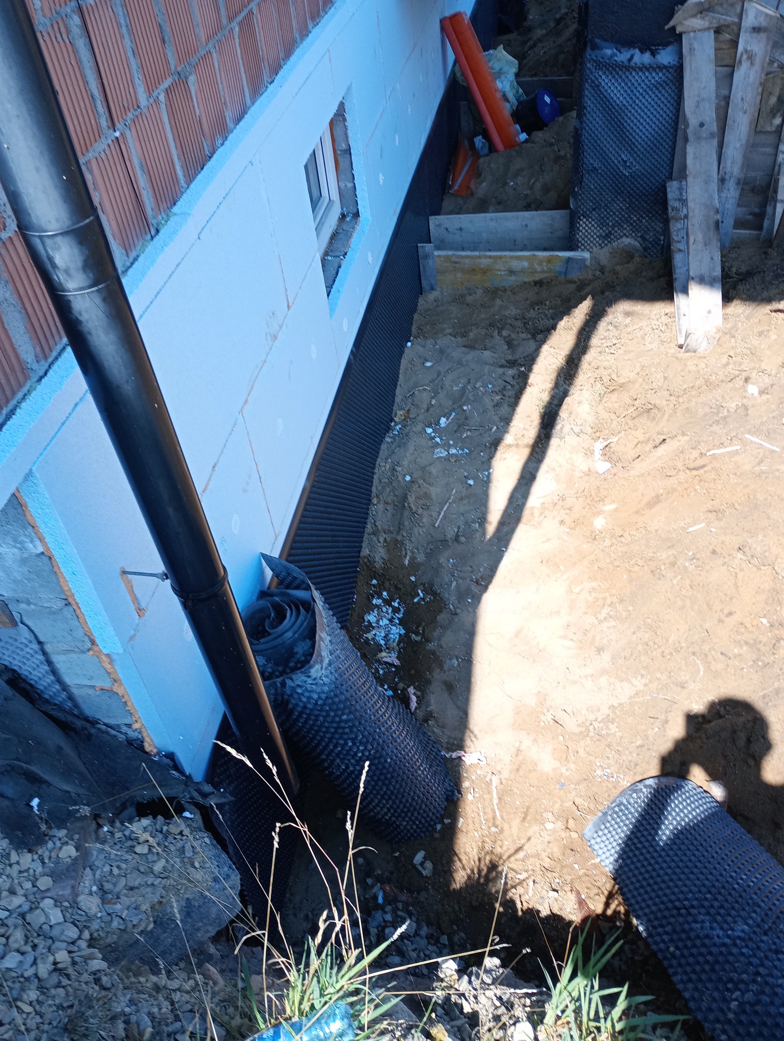 Ocieplenie fundamentów budynku w trakcie prac. Widoczna izolacja termiczna, folia kubełkowa i fragment ściany z cegły. Rury i elementy szalunkowe w wykopie.